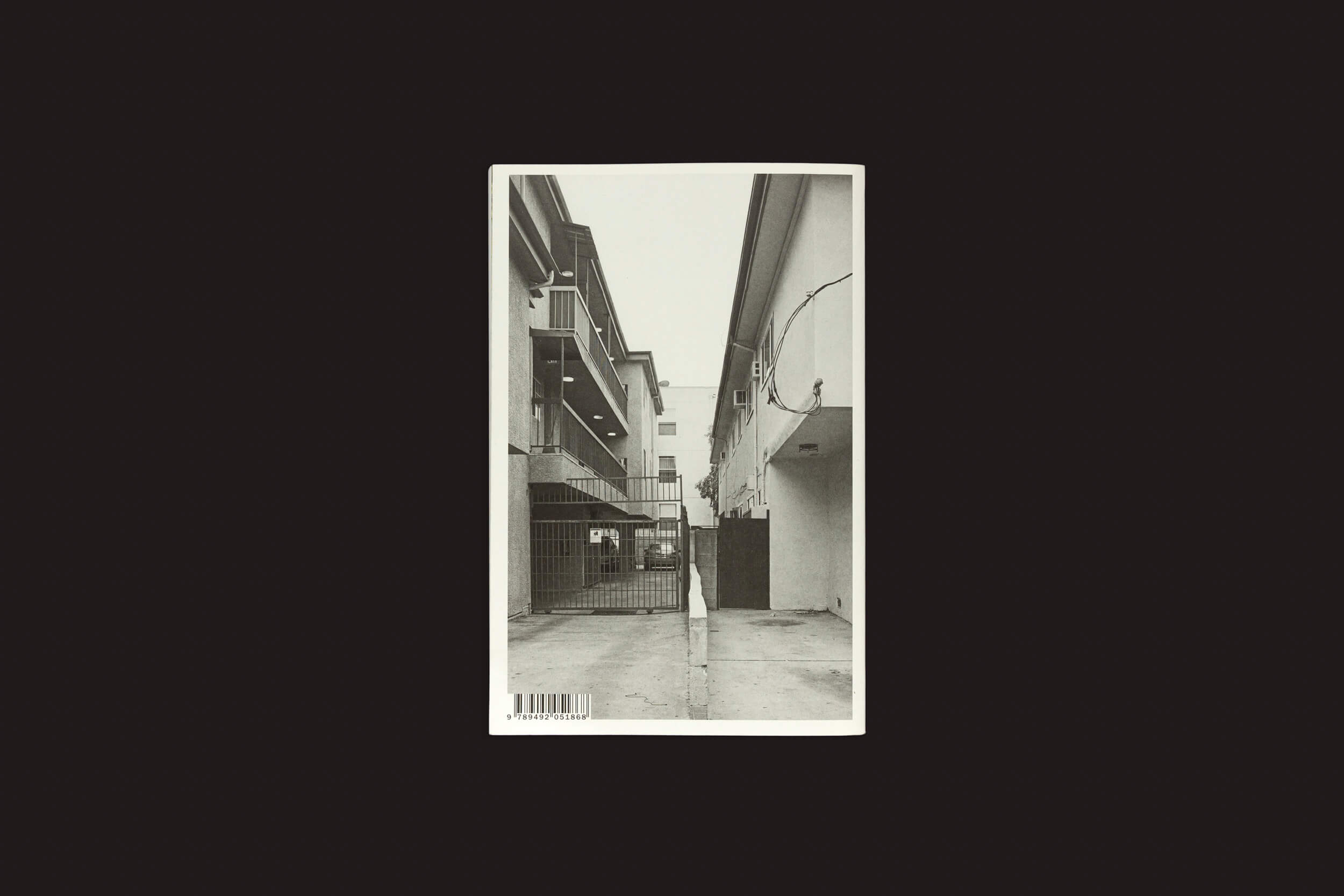 A foto em preto e branco de Thomas Locke Hobbs: L.A. Vedute, da The Eriskay Connection, captura um beco minimalista com varandas e um portão, exibindo simetria e perspectiva linear, que lembra a arquitetura doméstica.