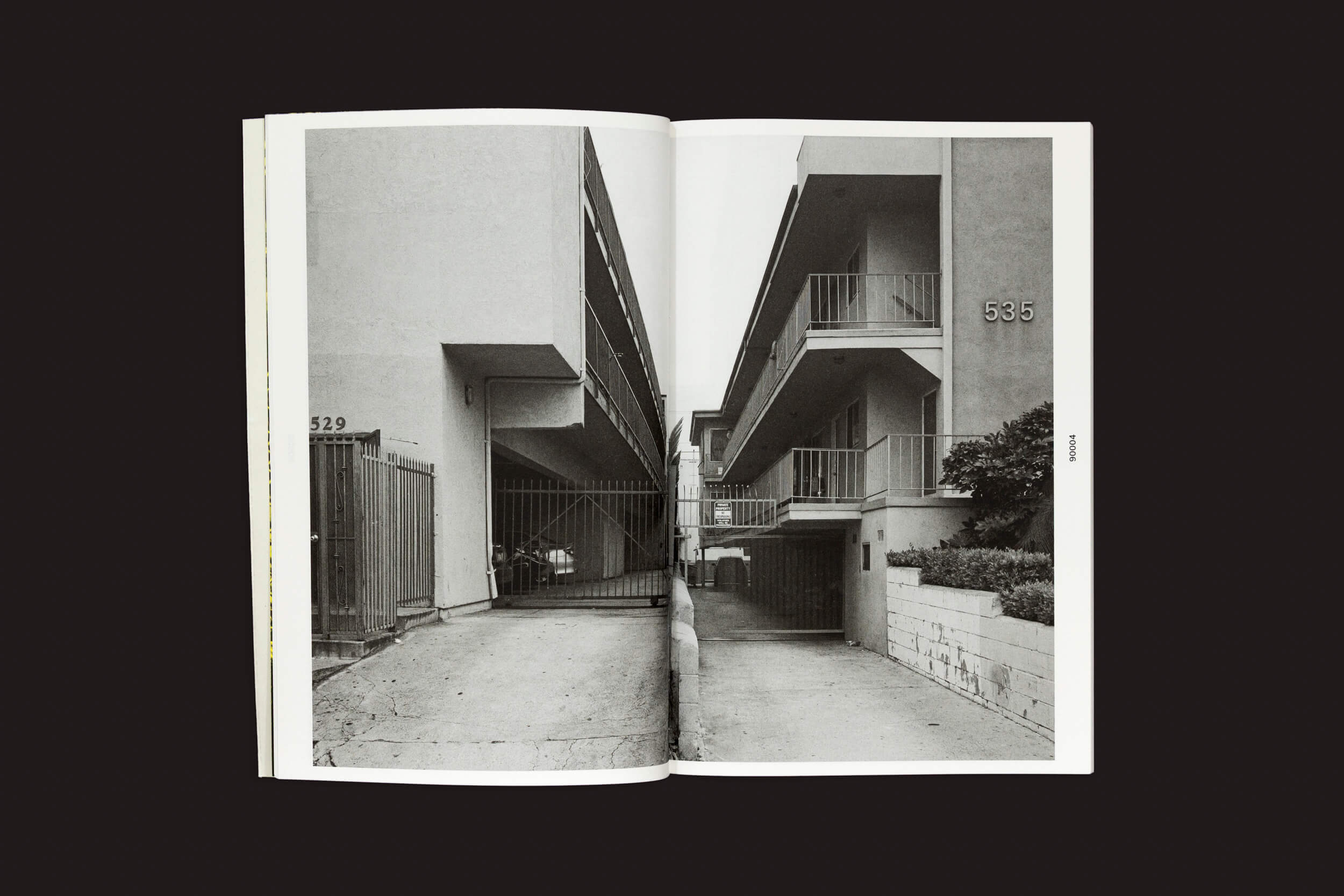 Um livro aberto, Thomas Locke Hobbs: L.A. Vedute, de The Eriskay Connection, apresenta uma impressionante fotografia em preto e branco da arquitetura doméstica dos edifícios adjacentes 529 e 535. Suas varandas e entrada com portão se destacam contra o fundo escuro.