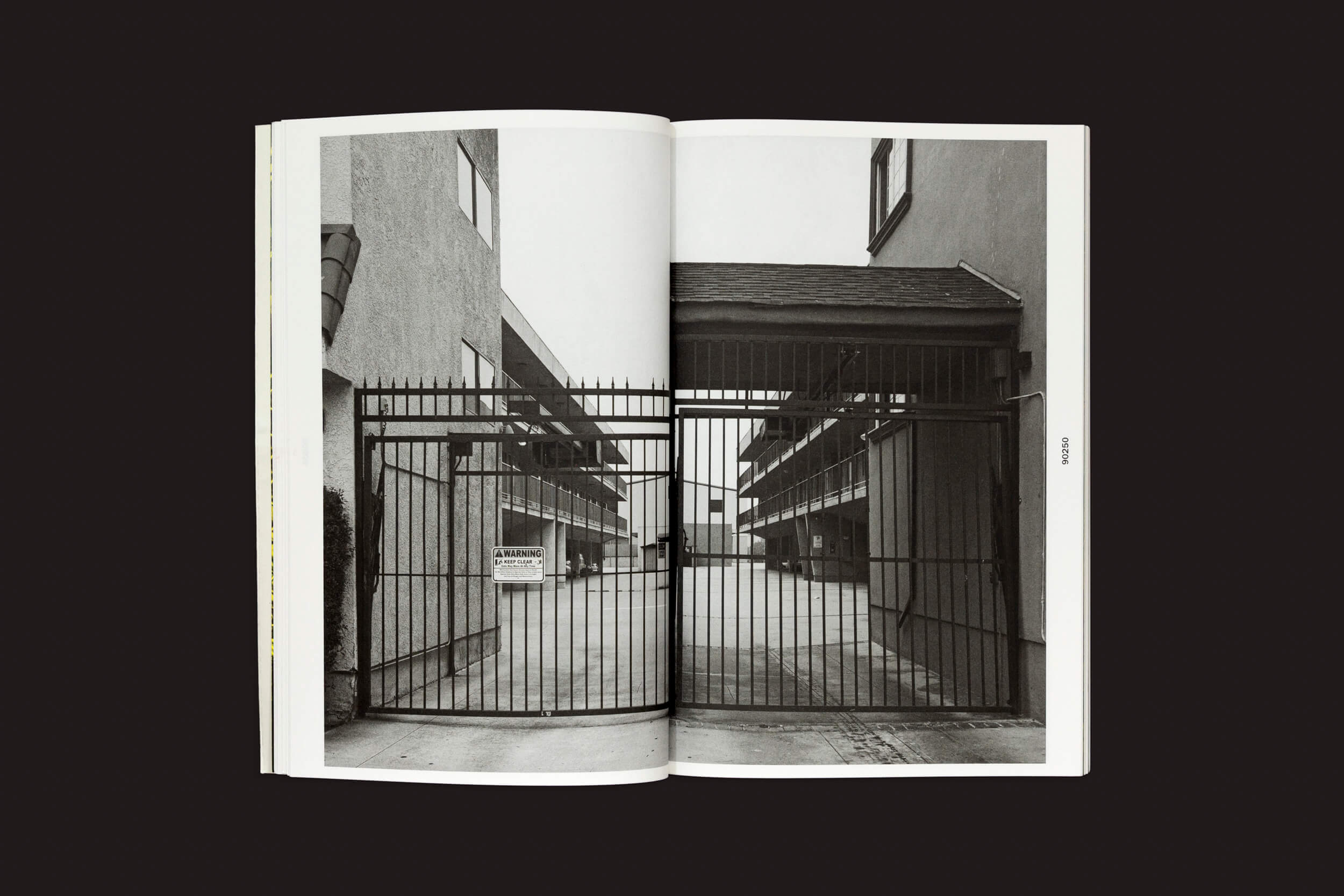 O livro aberto de Thomas Locke Hobbs: L.A. Vedute, do The Eriskay Connection, apresenta uma foto em preto e branco da entrada de um prédio de vários andares com portão, barras verticais e um interfone, encapsulando a essência minimalista e moderna da arquitetura doméstica.