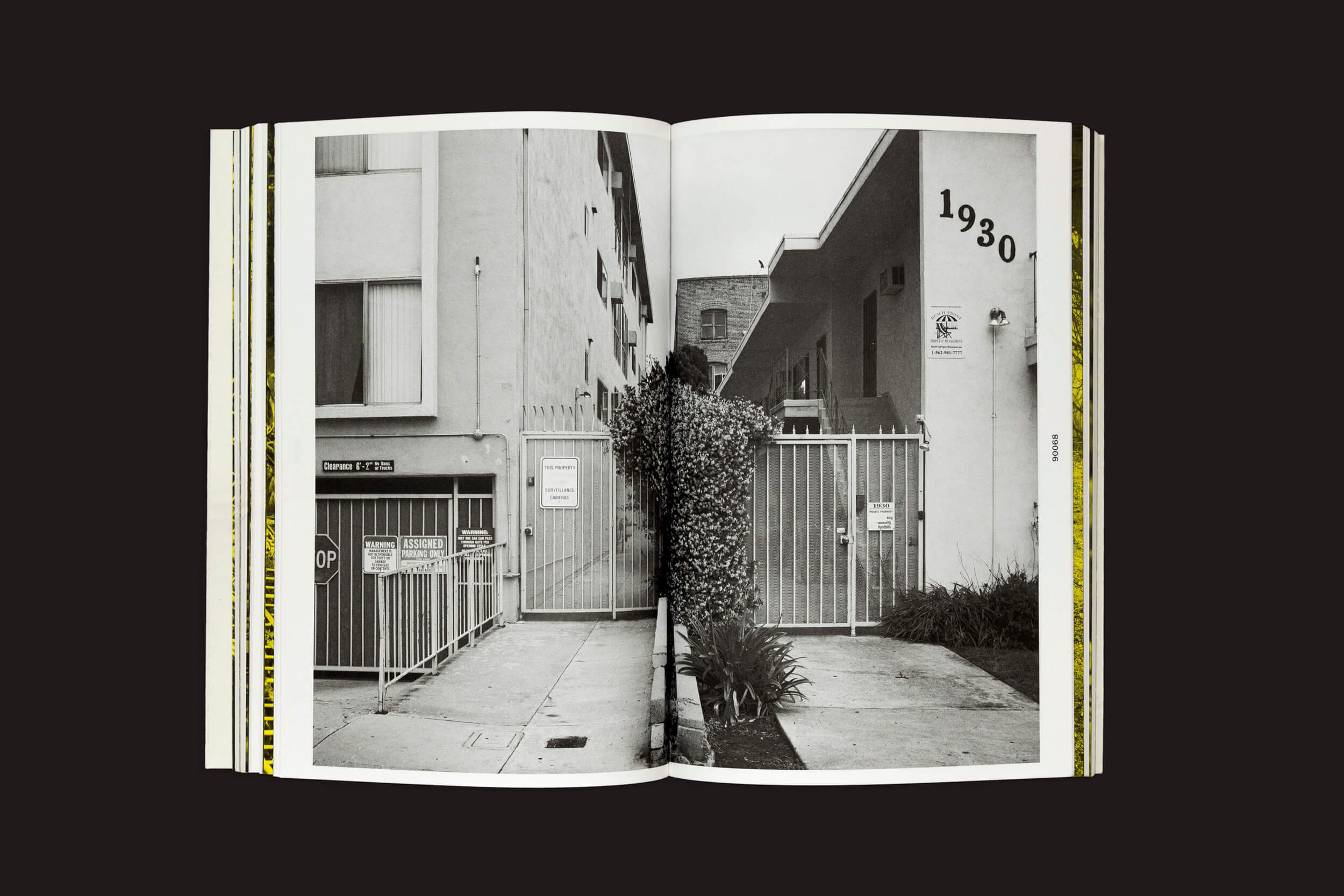 Um livro aberto de Thomas Locke Hobbs: L.A. Vedute do The Eriskay Connection revela uma foto em preto e branco de dois prédios divididos por uma cerca coberta de trepadeiras. O prédio da direita exibe 1930 e vários sinais, incorporando a arquitetura doméstica.