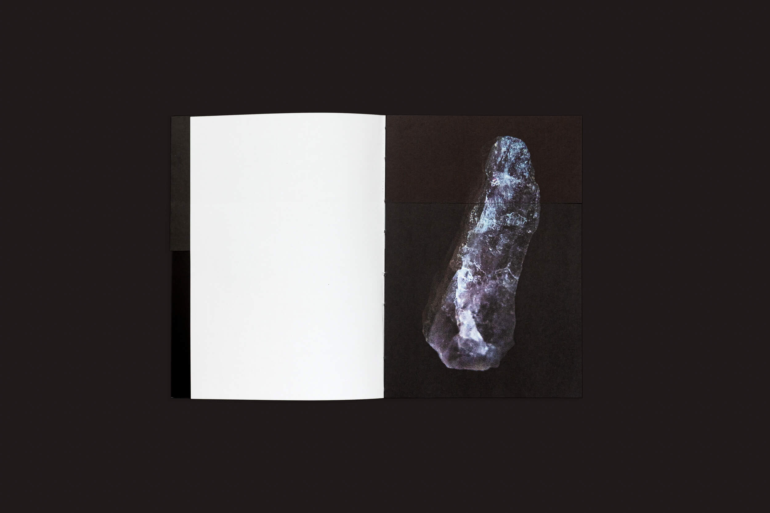 Uma cópia de Claudio Albarrán Briso: La herida del hombre da SED Editorial está aberta sobre uma superfície preta. A página esquerda em branco contrasta com as cores vibrantes de um longo cristal azul-púrpura à direita, convidando à introspecção contra a página totalmente branca e o fundo escuro.