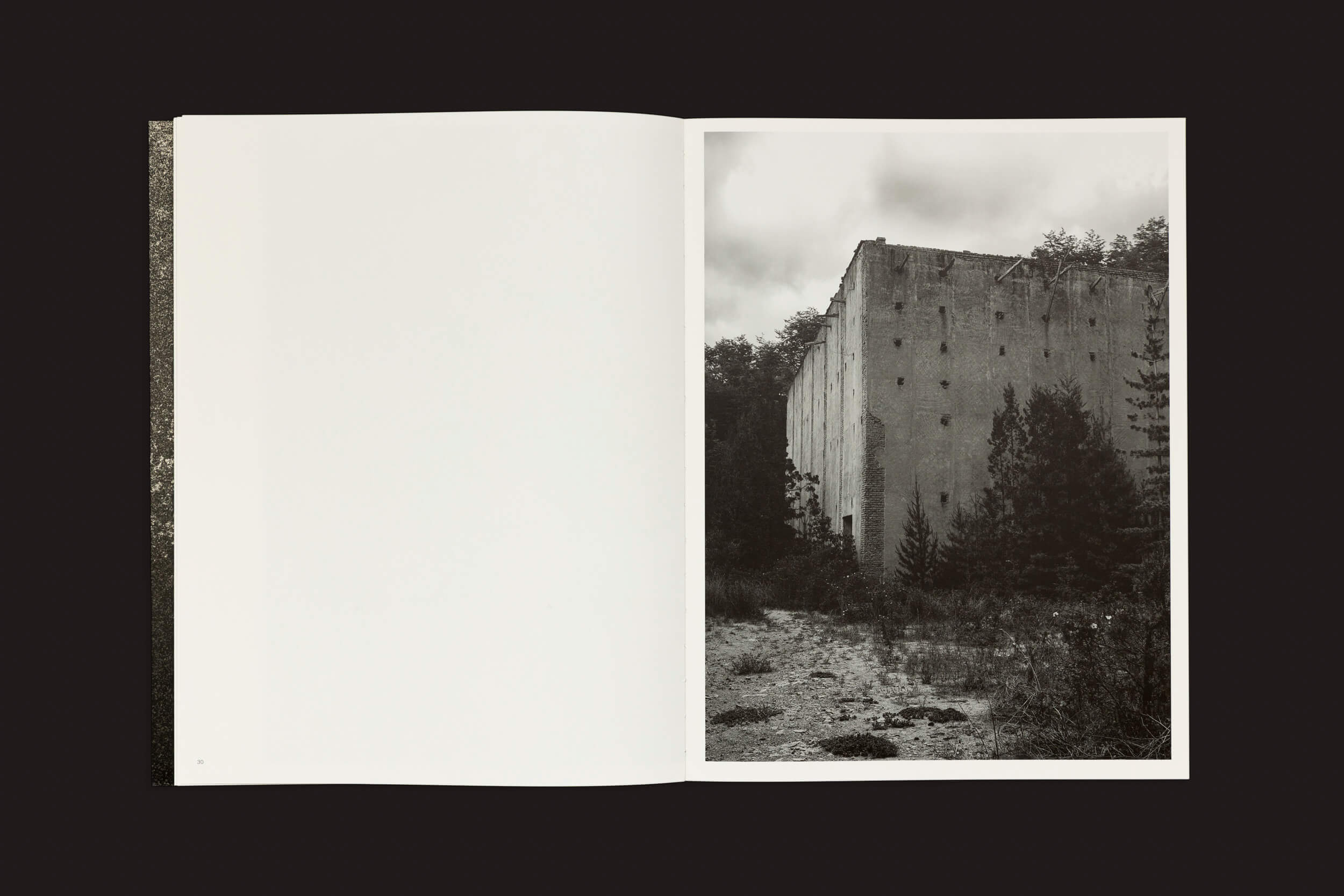 O livro aberto Pablo Cabado: Little Suns on Earth de The Eriskay Connection mostra uma foto em preto e branco de um prédio alto e retangular com muitas janelas, cercado por árvores e grama na Ilha Huemul. A página da esquerda permanece em branco.