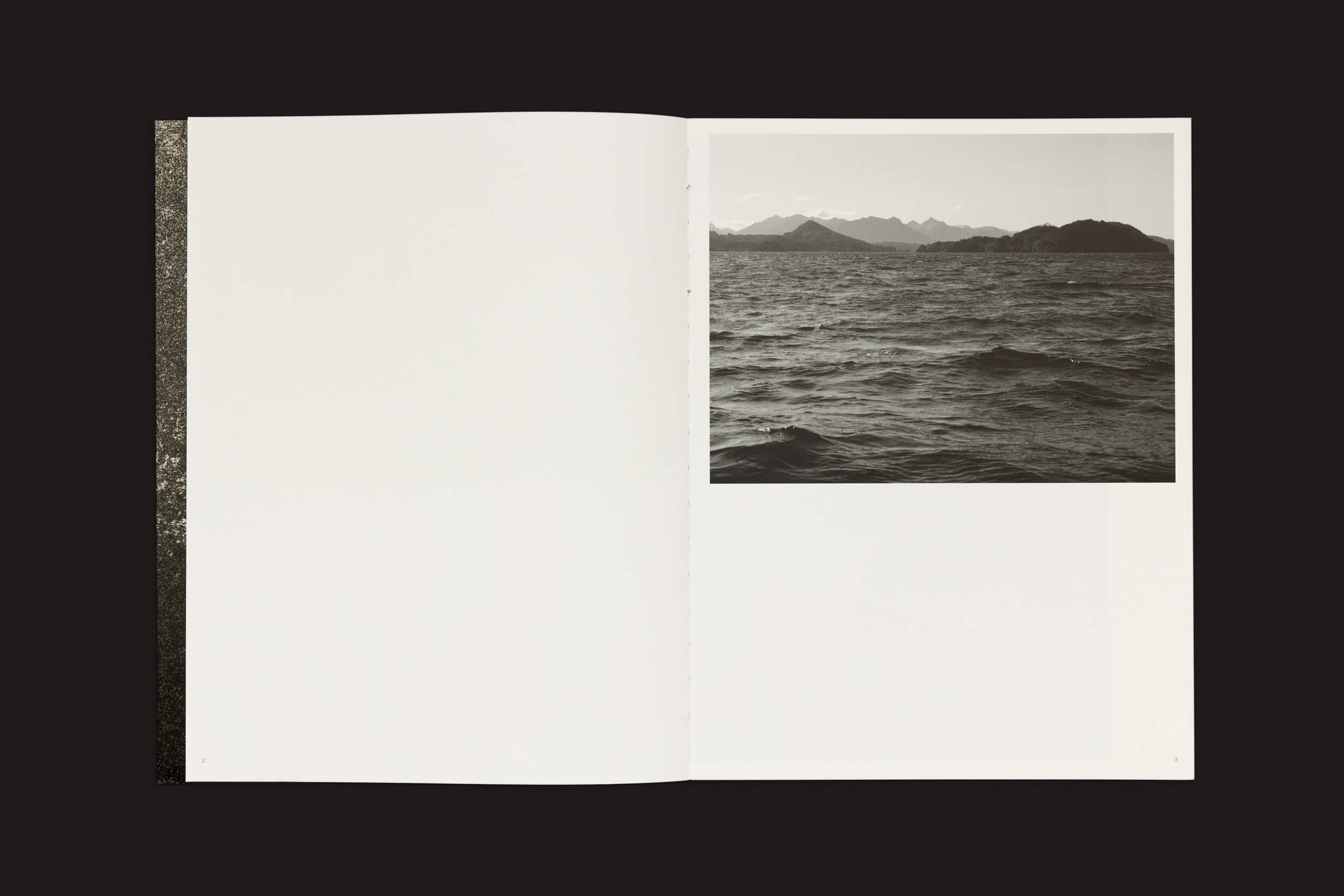 Em uma superfície escura, uma cópia aberta de Pablo Cabado: Little Suns on Earth, de The Eriskay Connection, revela uma foto em preto e branco de uma paisagem marinha na página direita, retratando ondas calmas e montanhas distantes sob um céu nublado que lembra a Ilha Huemul, enquanto a página esquerda está em branco.