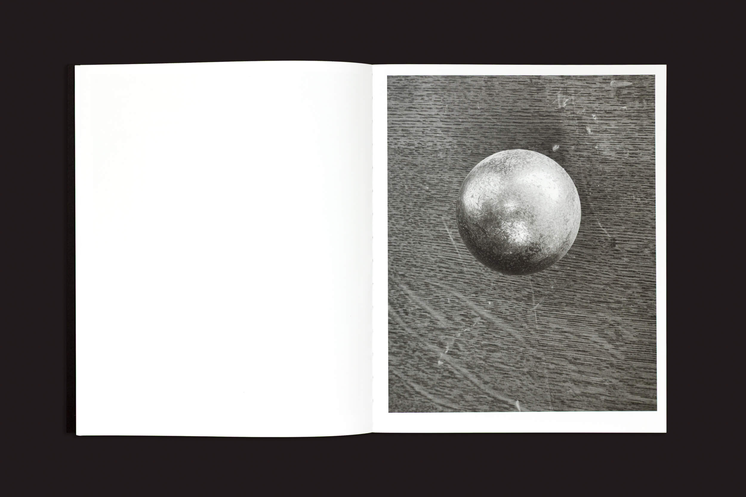 Um livro aberto intitulado "Ezio D'Agostino: NEOS", de Skinnerboox, apresenta uma foto em preto e branco de uma esfera metálica flutuando contra um fundo texturizado na página direita, enquanto a página esquerda permanece em branco.