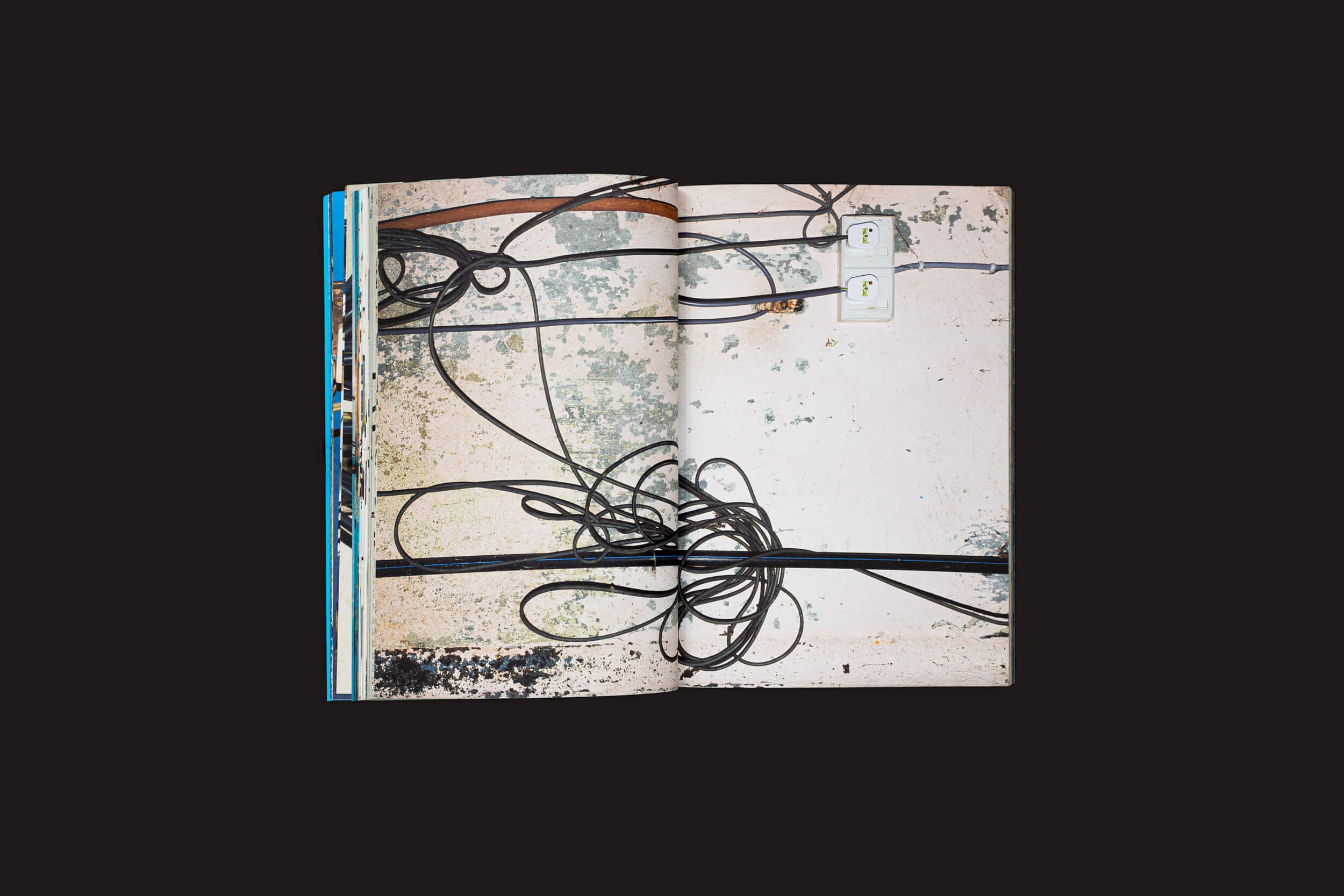 Henrik Malmström: NO da SED Editorial apresenta uma fotografia urbana de fios pretos emaranhados contra uma parede texturizada com duas tomadas brancas. As páginas se destacam contra um fundo preto, enfatizando as intervenções elétricas capturadas.