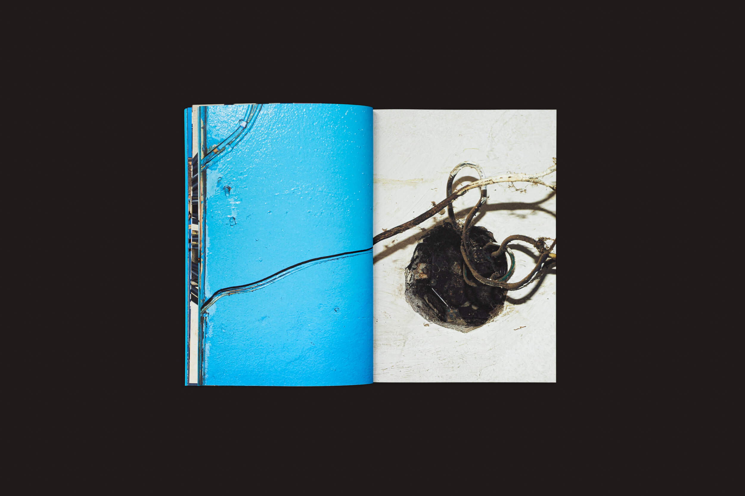 Uma revista aberta em um fundo preto revela Henrik Malmström: NO por SED Editorial. A página da esquerda apresenta um close-up de uma parede azul com uma rachadura preta, enquanto a da direita exibe uma composição abstrata de uma parede branca com fiação visível.