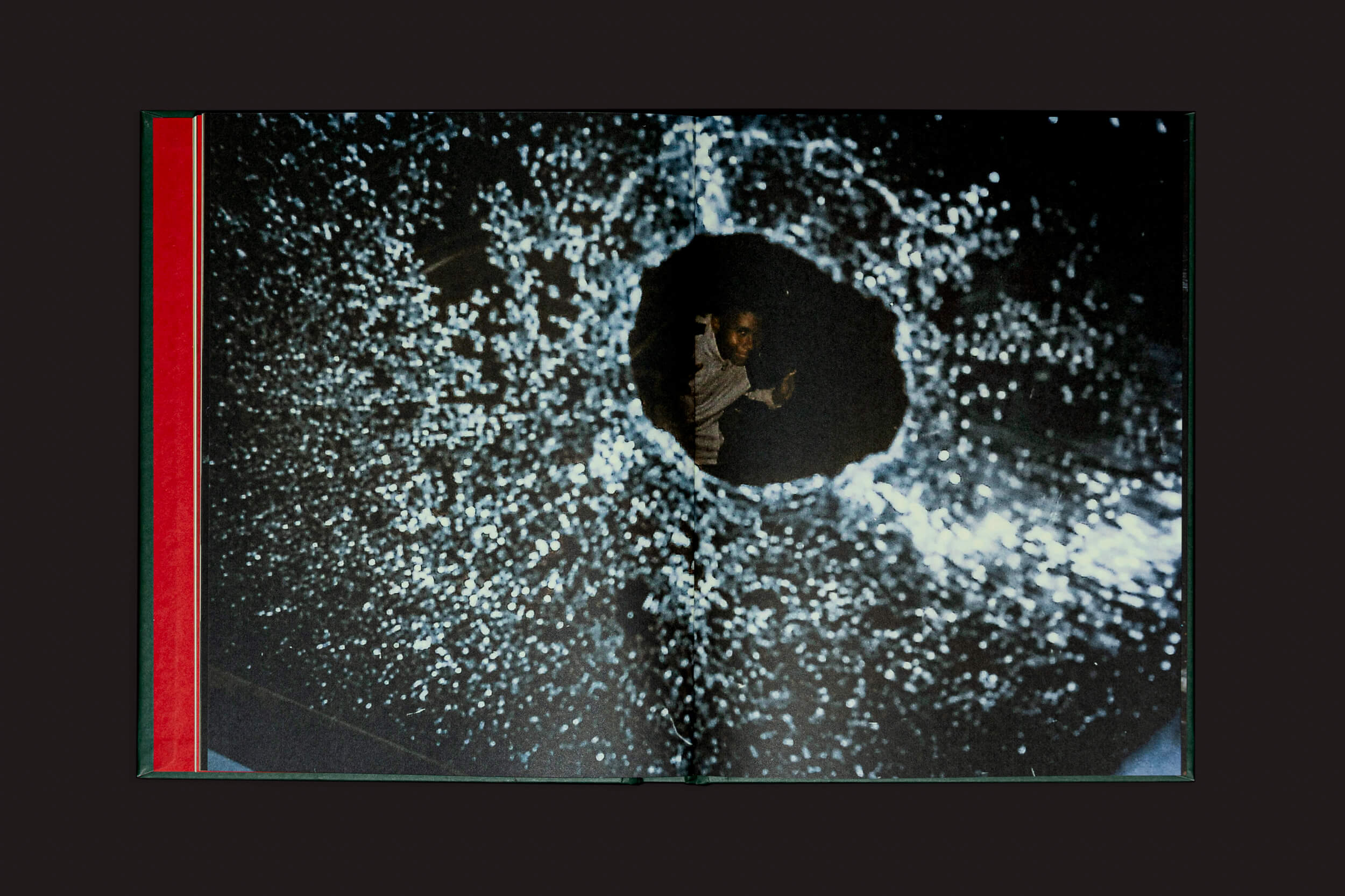 Um homem espia através de uma janela de vidro quebrada com um grande buraco circular, evocando uma memória familiar comovente. Esta cena noturna lembra Erick Peres: O choro pode durar uma noite, mas a alegria vem pela manhã, de Selo Turvo, destacando intrincados padrões de danos no vidro.