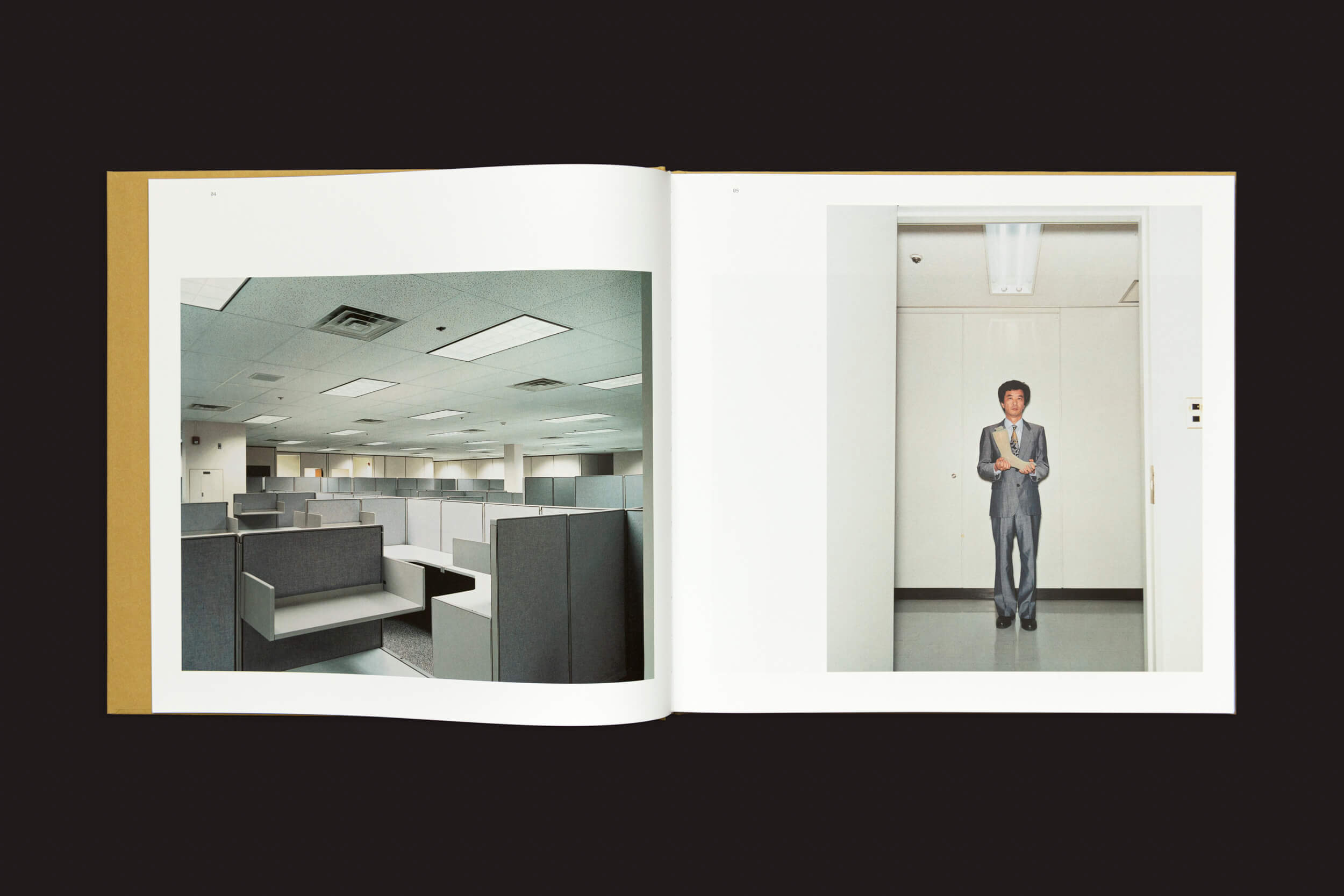 Um livro aberto, Lars Tunbjörk: Office / LA Office, de Loose Joints, mostra duas fotografias: a página da esquerda com um escritório deserto com cubículos cinzas e luzes de teto, e a página da direita capturando o estilo de Tunbjörk com uma pessoa de terno em um corredor de paredes brancas sob luzes de teto.