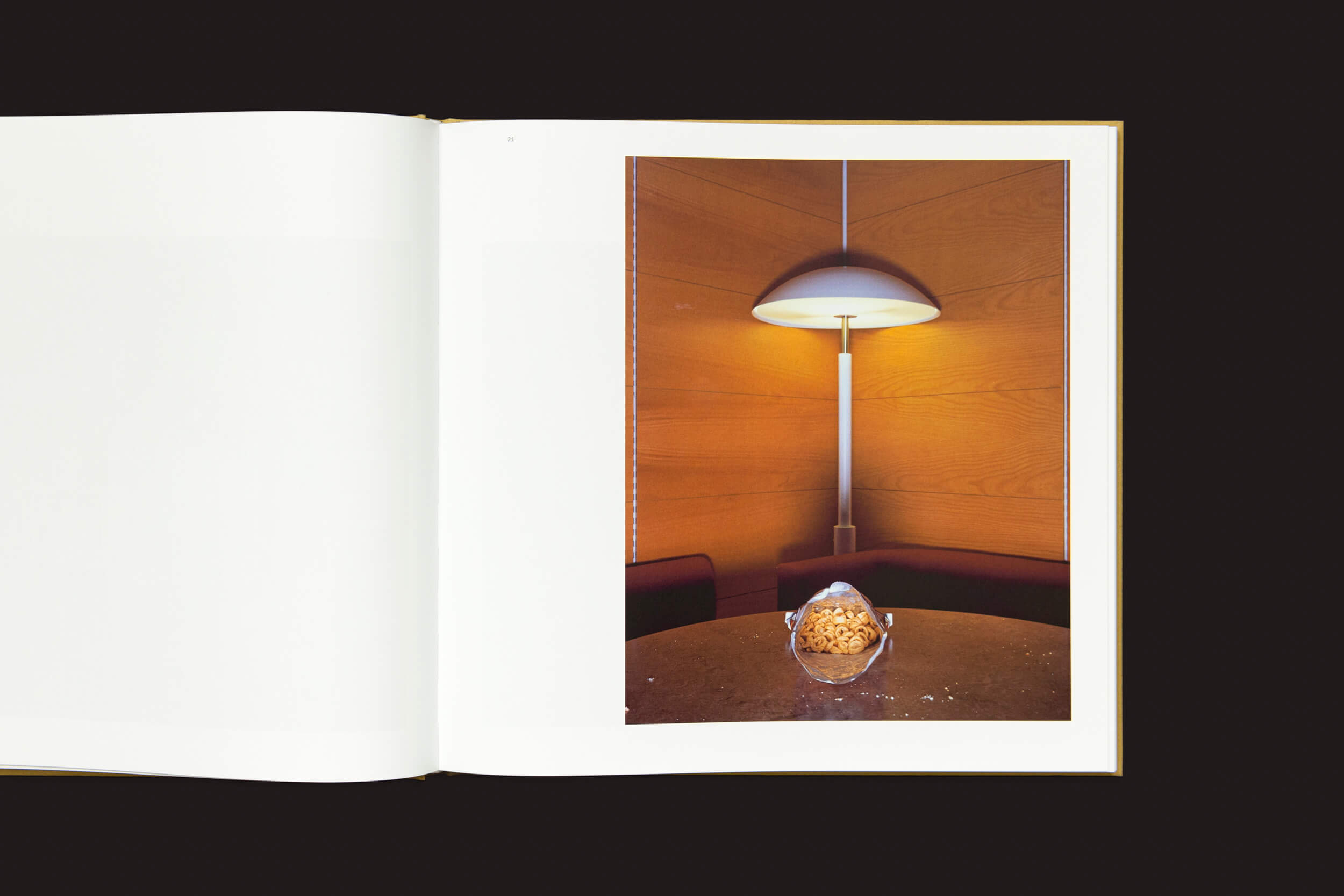 Um livro aberto revela uma fotografia moderna de interior com paredes de madeira, uma mesa redonda, luminária de teto e peça central de vidro decorativa — uma cena que evoca Office / LA Office de Lars Tunbjörk por Loose Joints. A página oposta está em branco, capturando a essência do trabalho remoto.