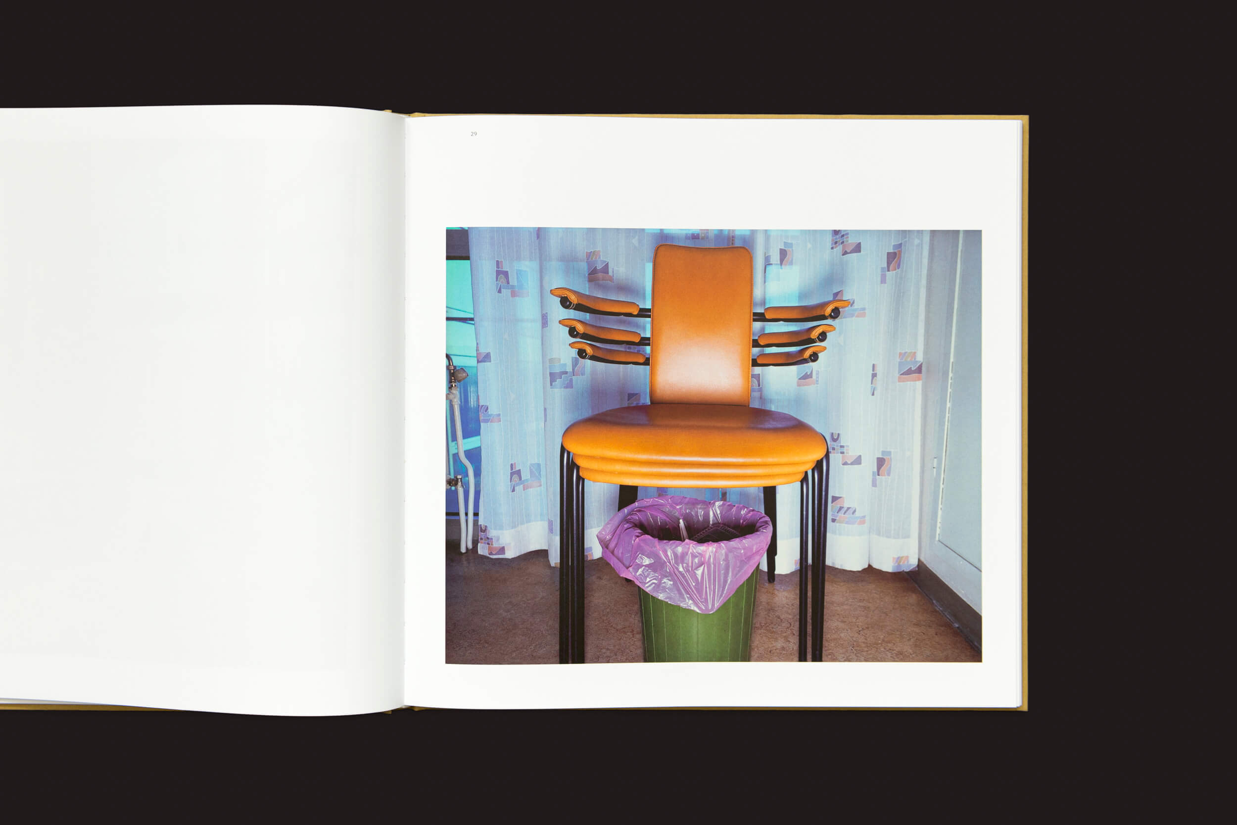 Um livro aberto apresenta uma fotografia de Lars Tunbjörk: Office / LA Office, da Loose Joints: três cadeiras laranja empilhadas em uma lixeira verde com um forro roxo, contra cortinas com padrões claros e um fundo escuro.