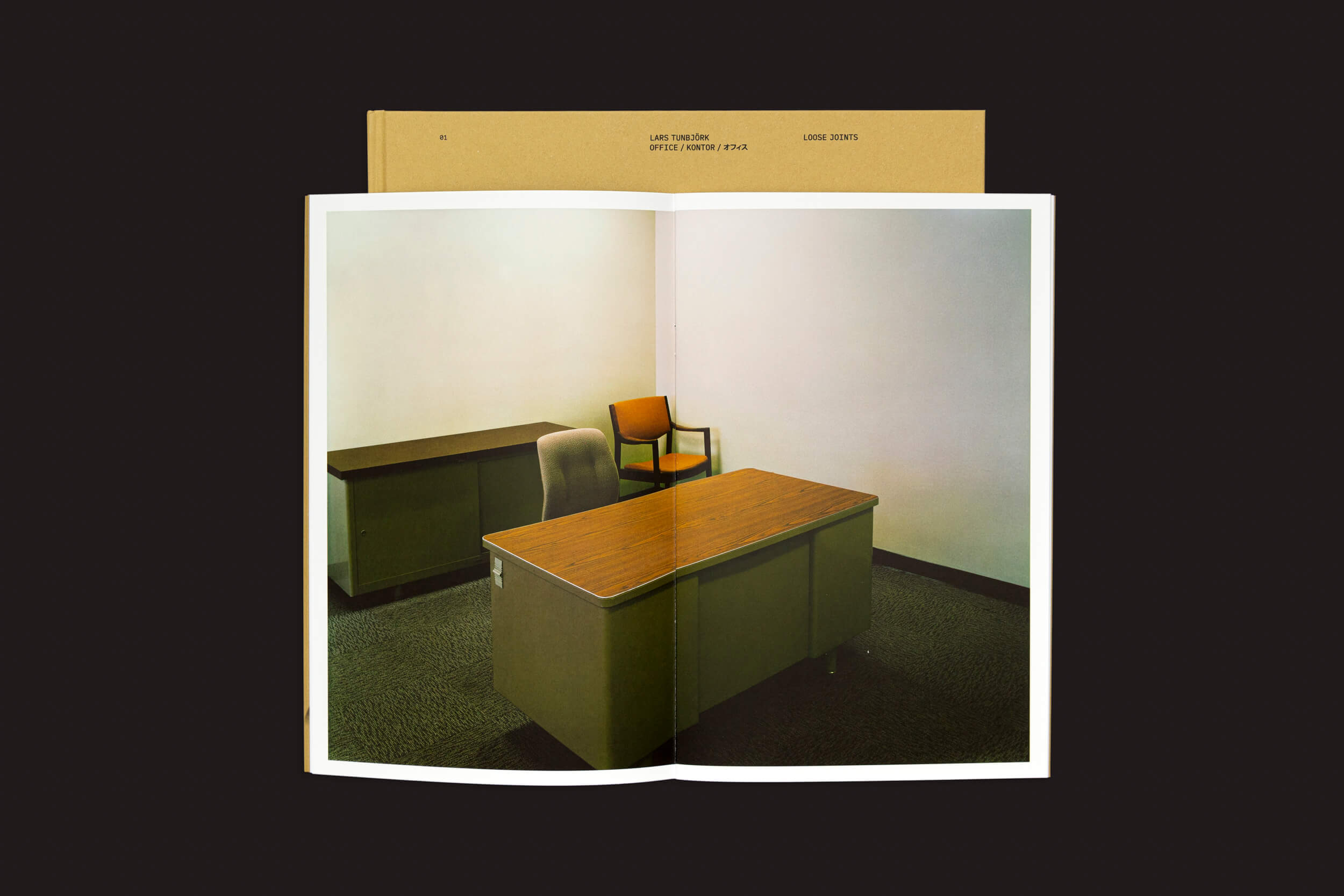 Um livro aberto, Lars Tunbjörk: Office / LA Office de Loose Joints, mostra uma cena de escritório vazia com uma mesa e cadeira de madeira, ecoando o estilo de Tunbjörk. Uma cadeira laranja fica no canto contra uma parede simples, sugerindo uma mudança para o trabalho remoto. A capa do livro é parcialmente visível atrás das páginas.