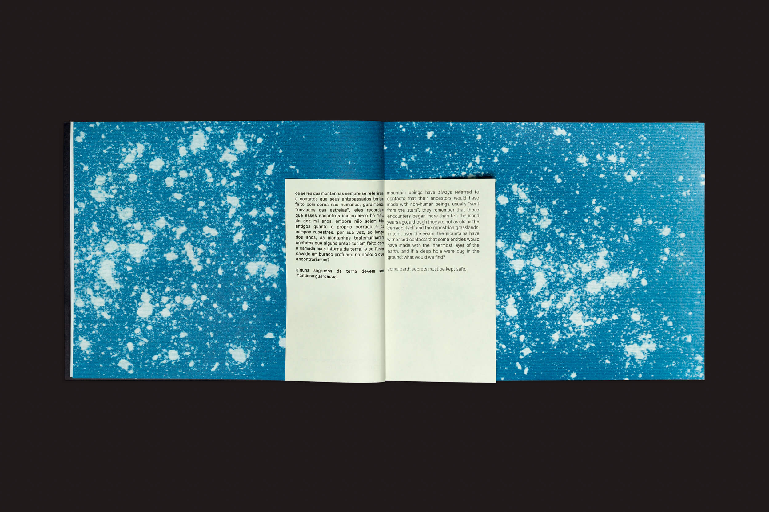 Um livro com uma página de texto central é colocado contra um fundo azul com respingos brancos. O bloco de texto direito está alinhado, o esquerdo não, em uma sobreposição bege que lembra imagens-paisagens da Info Óris. Inspirado por Óris de Bárbara Lissa e Maria Vaz do Selo Turvo.