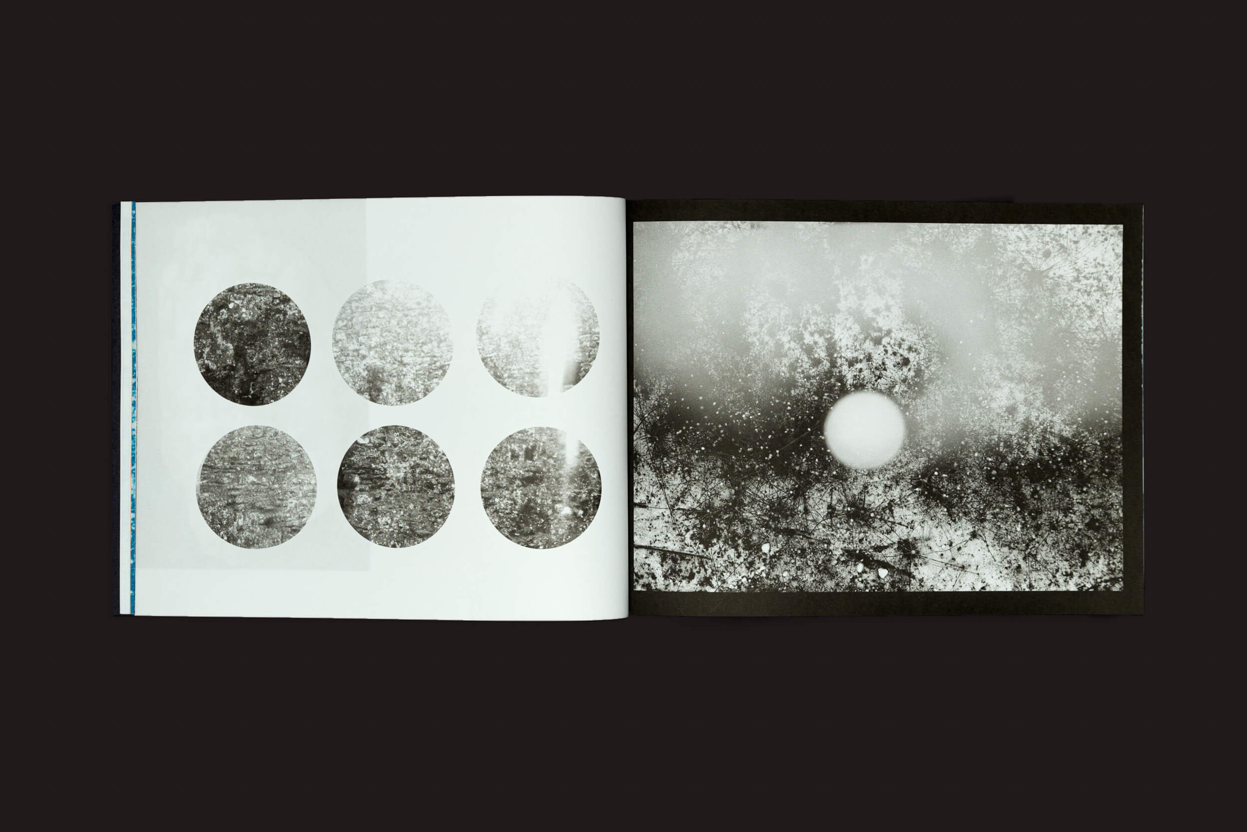 Um livro aberto sobre uma superfície escura apresenta elementos de Bárbara Lissa & Maria Vaz: Óris de Selo Turvo. A página da esquerda mostra seis círculos texturizados em duas fileiras, e a da direita exibe uma imagem abstrata monocromática com um grande círculo branco e fundo salpicado, evocando temas do Antropoceno.