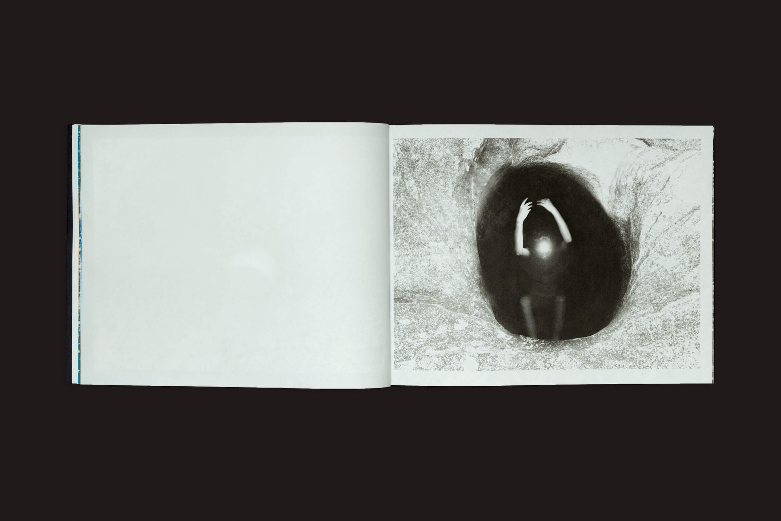 Um livro aberto intitulado Óris de Bárbara Lissa & Maria Vaz do Selo Turvo revela uma imagem em preto e branco na página direita, que lembra temas do Antropoceno, com uma figura emergindo de uma forma oval escura, com as mãos estendidas para cima. A página esquerda em branco convida a interpretações por meio do contraste.