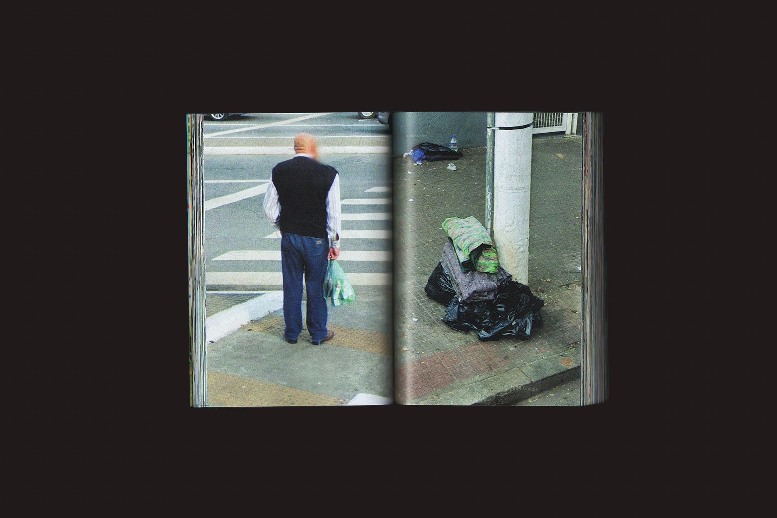 Uma foto de "Romeu Silva: Passantes", da Zero-Edições, captura um homem idoso e careca em uma faixa de pedestres de São Paulo com sacolas plásticas, enquanto, ao lado, pertences embrulhados em cobertores e sacolas descansam em um poste na calçada.