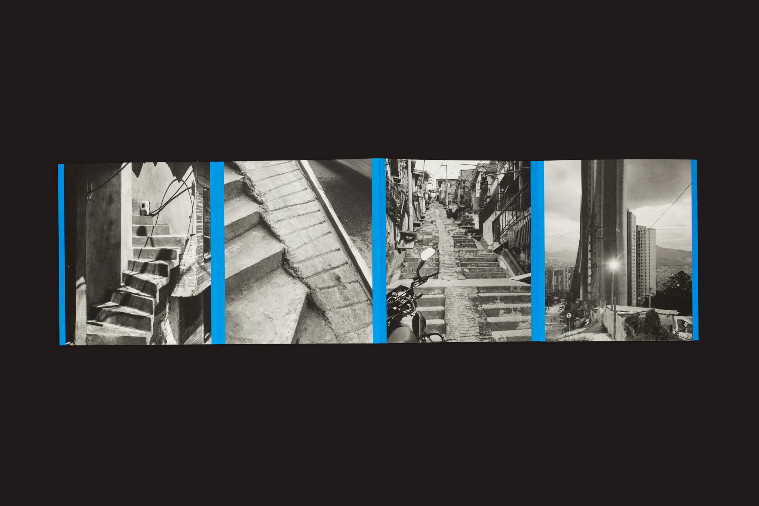 Rampitas de Thomas Locke Hobbs, da The Eriskay Connection, apresenta uma colagem de fotografias em preto e branco sobre um fundo preto, capturando a vida urbana com imagens de escadas de concreto, rampas esculturais, ruas estreitas com linhas de energia e estruturas imponentes separadas por elegantes linhas verticais azuis.