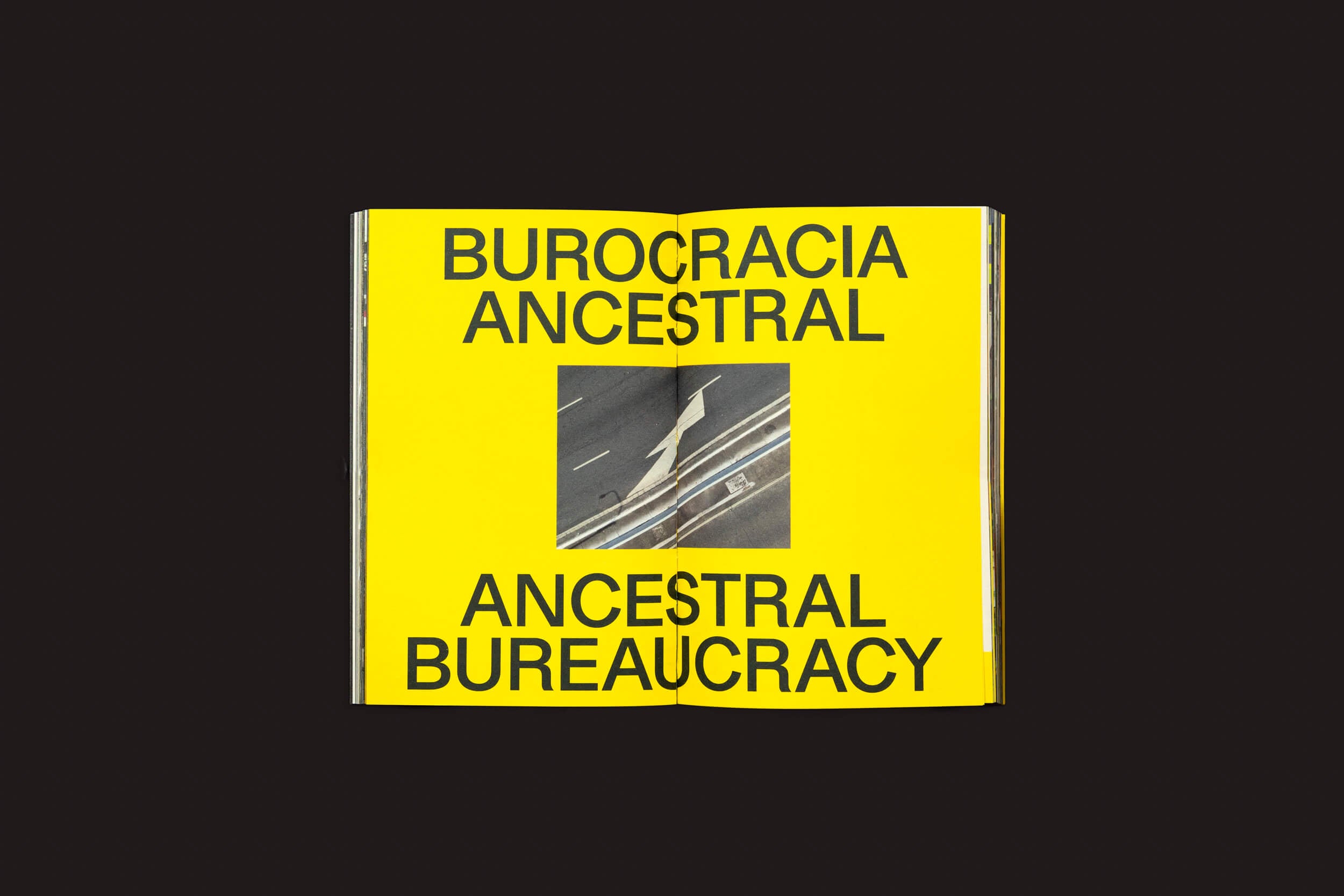 Um livro aberto da Livros Fantasma intitulado Ermãos Monjon: Sinais/Signs traz o texto em negrito e preto BUROCRACIA ANCESTRAL e BUROCRACIA ANCESTRAL em páginas amarelas. Aparece uma fotografia aérea de estrada que lembra deriva urbana, em um fundo preto que evoca misteriosos caminhos linguísticos.