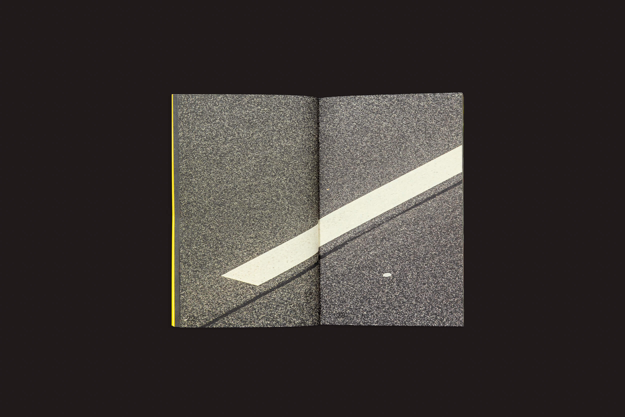 O livro Ermãos Monjon: Sinais/Signs da Livros Fantasma revela uma estrada de asfalto com uma linha diagonal branca e um ponto, ecoando o estilo intrigante de Ermãos Monjon. O fundo escuro acentua a textura, convidando à exploração de linguagens gráficas.