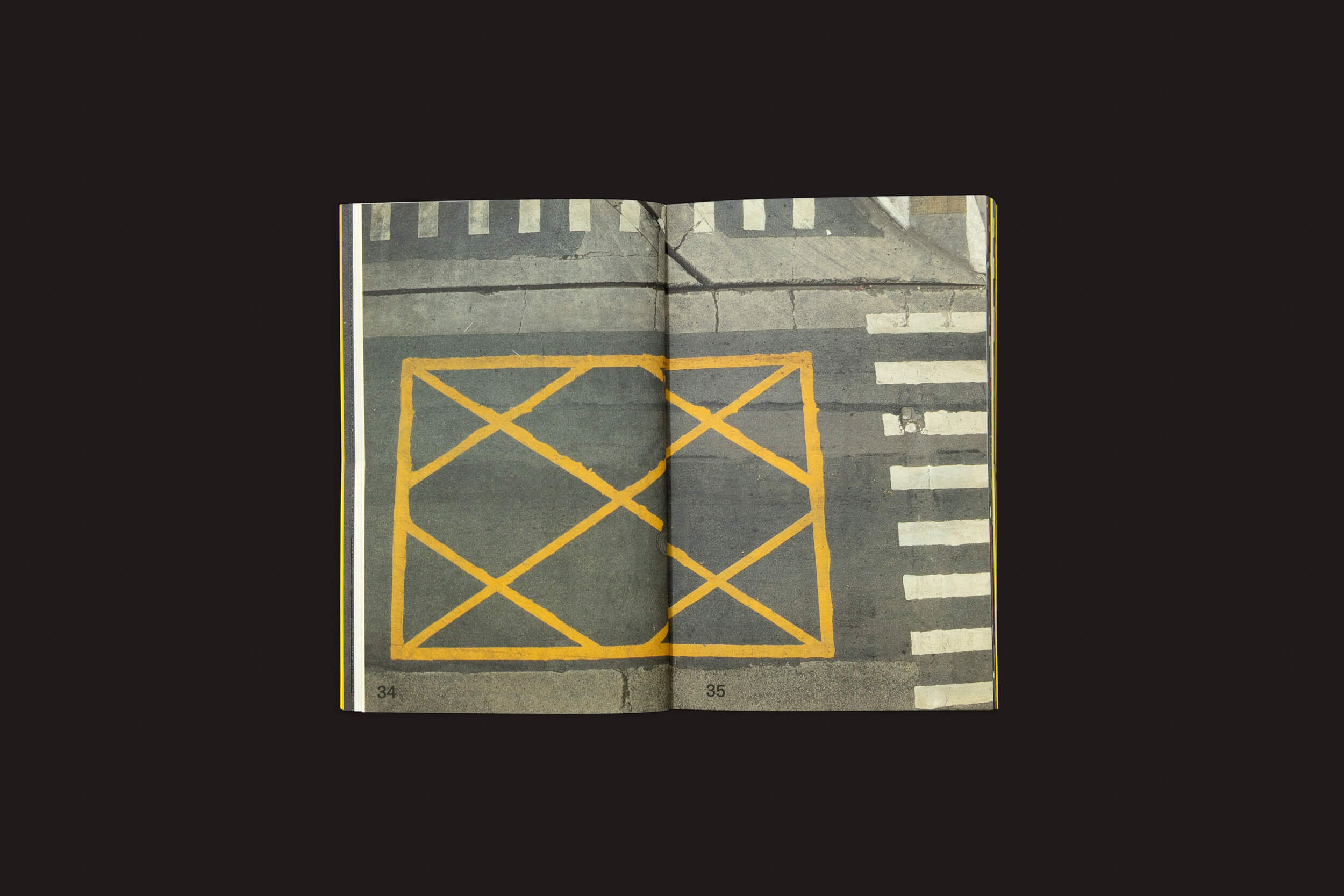 Uma revista aberta da Livros Fantasma, Ermãos Monjon: Sinais/Signs, revela uma foto de cruzamento de rua. Linguagens gráficas vívidas, linhas de faixa de pedestres e um padrão de grade amarela adornam a cena no verdadeiro estilo Ermãos Monjon. As páginas 34-35 estão claramente marcadas na parte inferior.