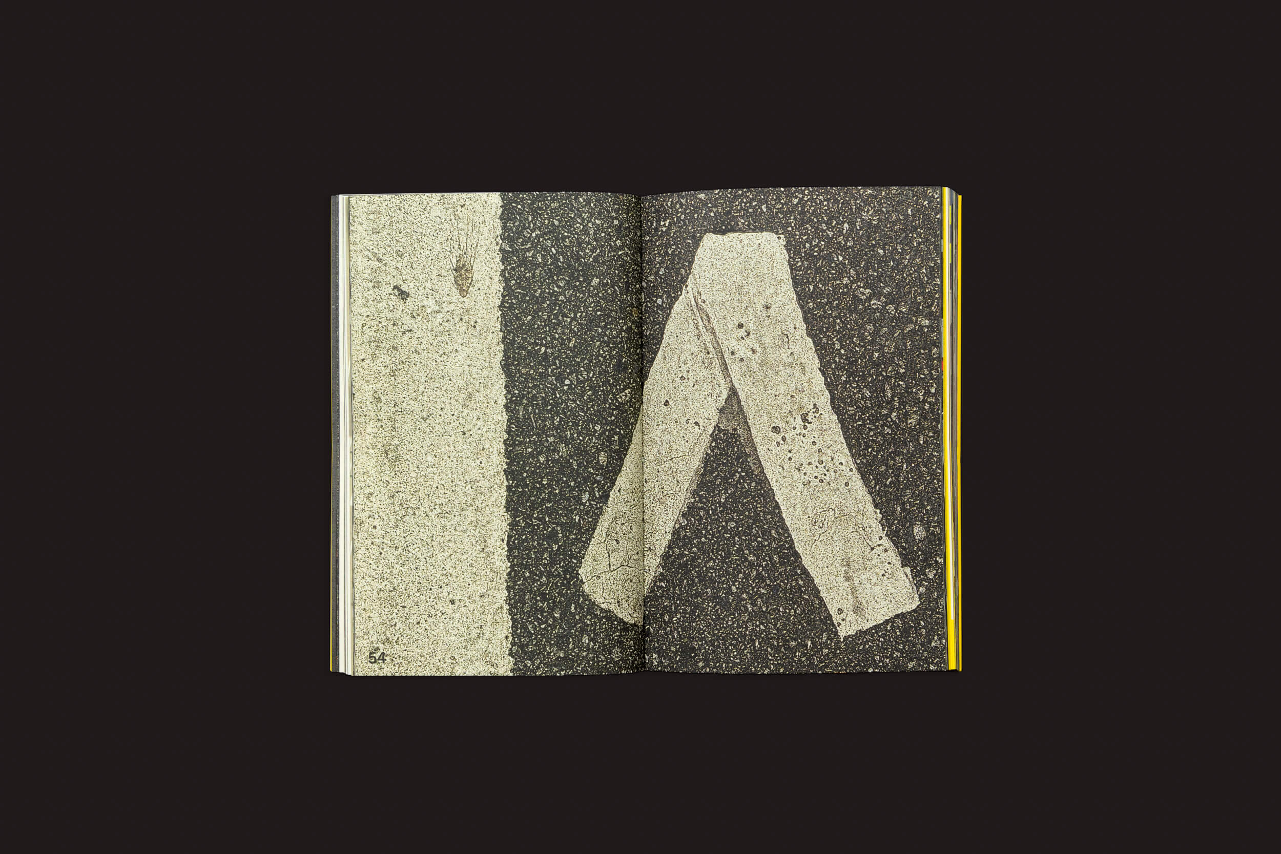 O livro aberto Ermãos Monjon: Sinais/Signs da Livros Fantasma revela uma foto de marcações rodoviárias em asfalto texturizado, apresentando uma linha branca reta em uma página e uma marcação em forma de chevron na outra. O fundo preto sólido ecoa uma peça de Ermãos Monjon, capturando a essência das linguagens gráficas.