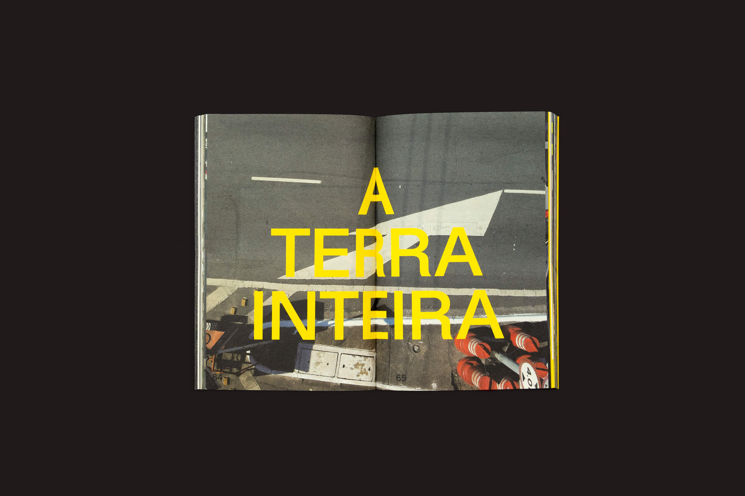 Um livro aberto retrata uma cena de rua com faixa de pedestres e cones de trânsito, capturando deriva urbana. Um grande texto amarelo dizendo A TERRA INTEIRA aparece contra o fundo preto, destacando as linguagens gráficas de Ermãos Monjon: Sinais/Signs da Livros Fantasma.
