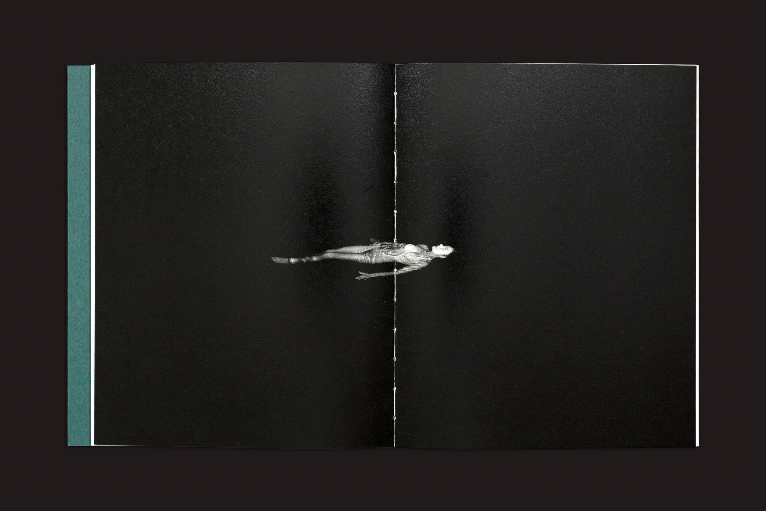 Uma página de Dylan Hausthor e Pia Paulina Guilmoth: Sleep Creek (Vazio) mostra uma pequena figura em tons de cinza flutuando horizontalmente no centro sobre um fundo preto, dividida verticalmente pelo vinco do livro.