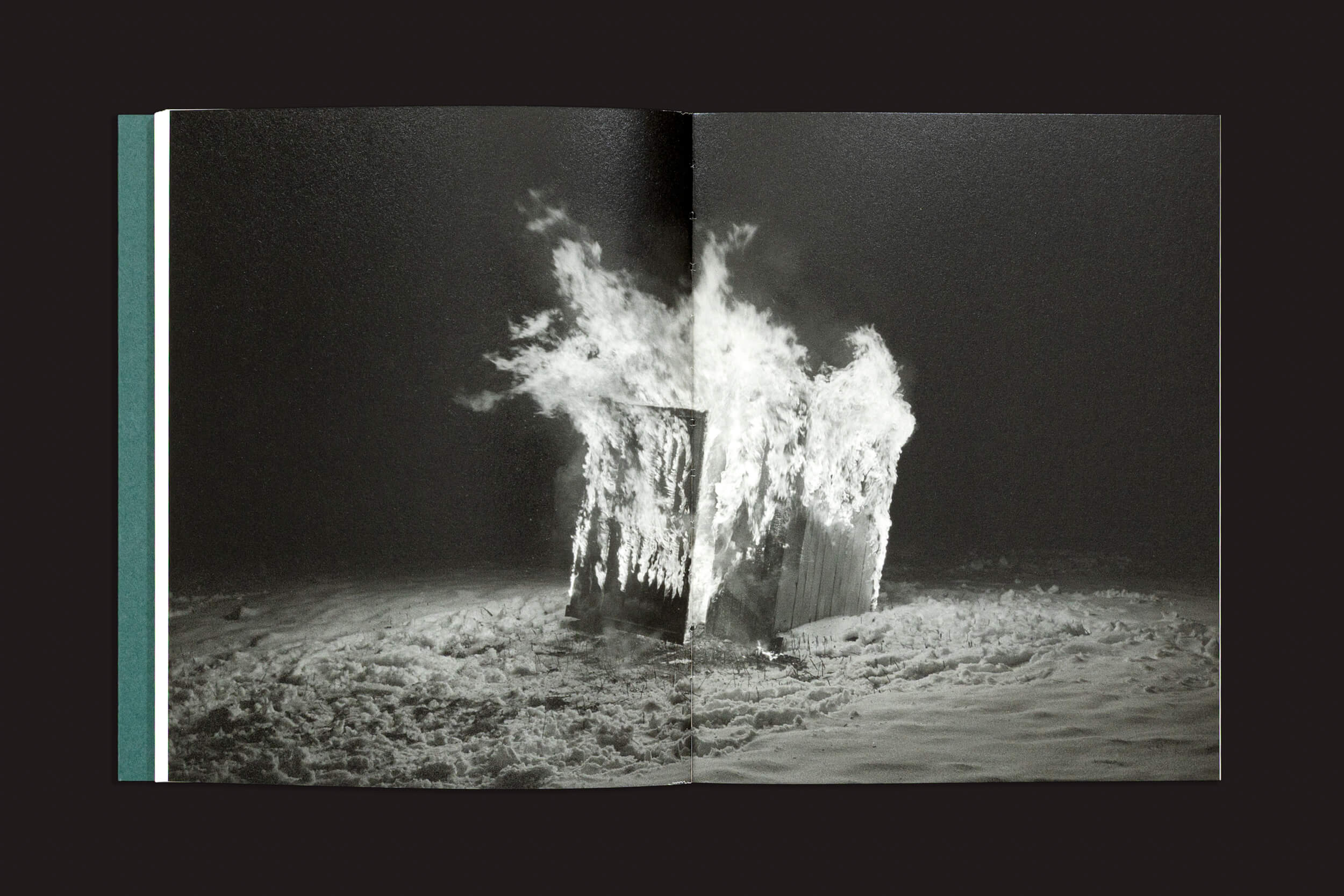 Uma fotografia em preto e branco no estilo da fotografia documental e ficcional mostra uma caixa de madeira em chamas na areia, com fumaça subindo contra a escuridão. Apresentada como uma página dupla em "Dylan Hausthor & Pia Paulina Guilmoth: Sleep Creek", da Void.