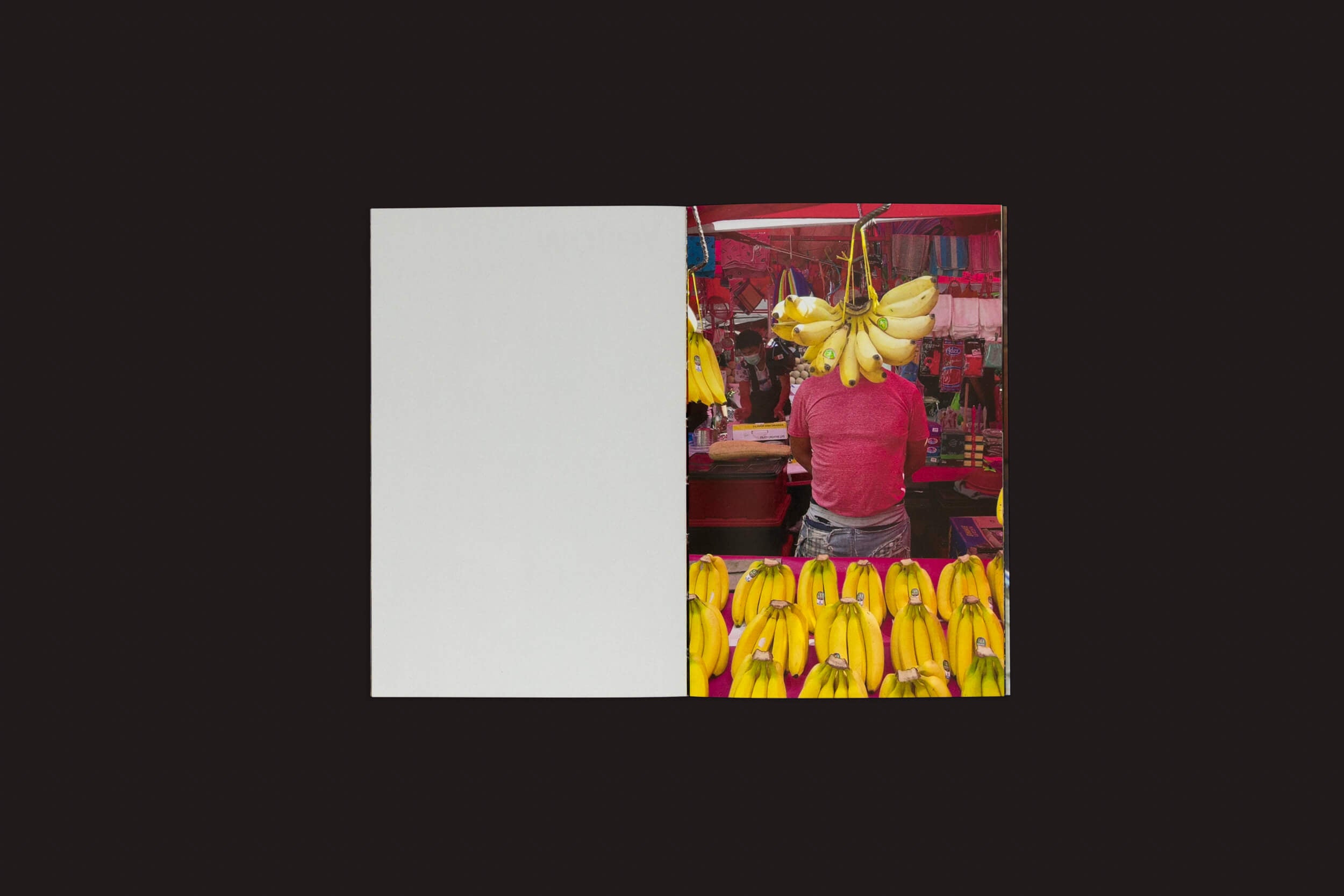 Uma pessoa de camisa vermelha está em uma barraca de frutas com cachos de bananas amarelas, capturando uma obra observacional do vibrante mercado. A imagem se divide, deixando o lado esquerdo em branco e enfatizando o movimentado lado direito — a essência da experiência humana em Sam Youkilis: Somewhere 2017-2023 de Loose Joints.