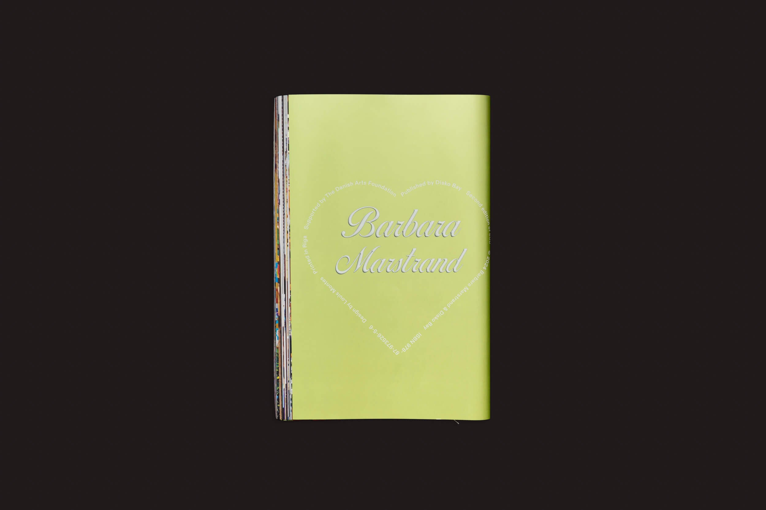 Um fotolivro fechado com capa verde contra um fundo preto. A capa exibe um coração branco com Barbara Marstrand dentro e um texto menor ao redor. Suas páginas multicoloridas refletem a vibrante cultura jovem dinamarquesa. Marca: Disko Bay.