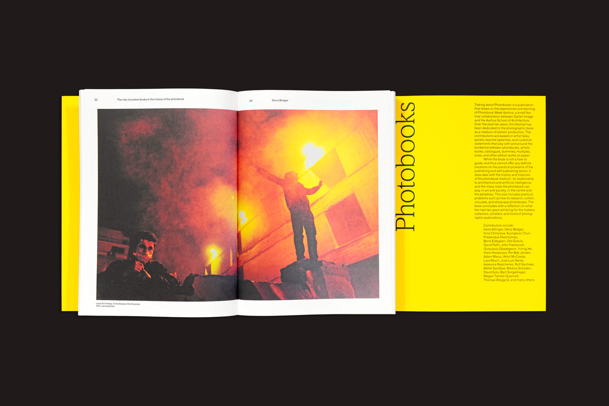 Um livro aberto em uma superfície escura mostra uma foto de duas pessoas com sinalizadores em meio à fumaça e à luz. Intitulado "Talking About Photobooks" por Moritz Neumüller, ele examina a história e o impacto dos fotolivros na arte e na sociedade, publicado pela Fw:Books com uma capa amarela vívida.