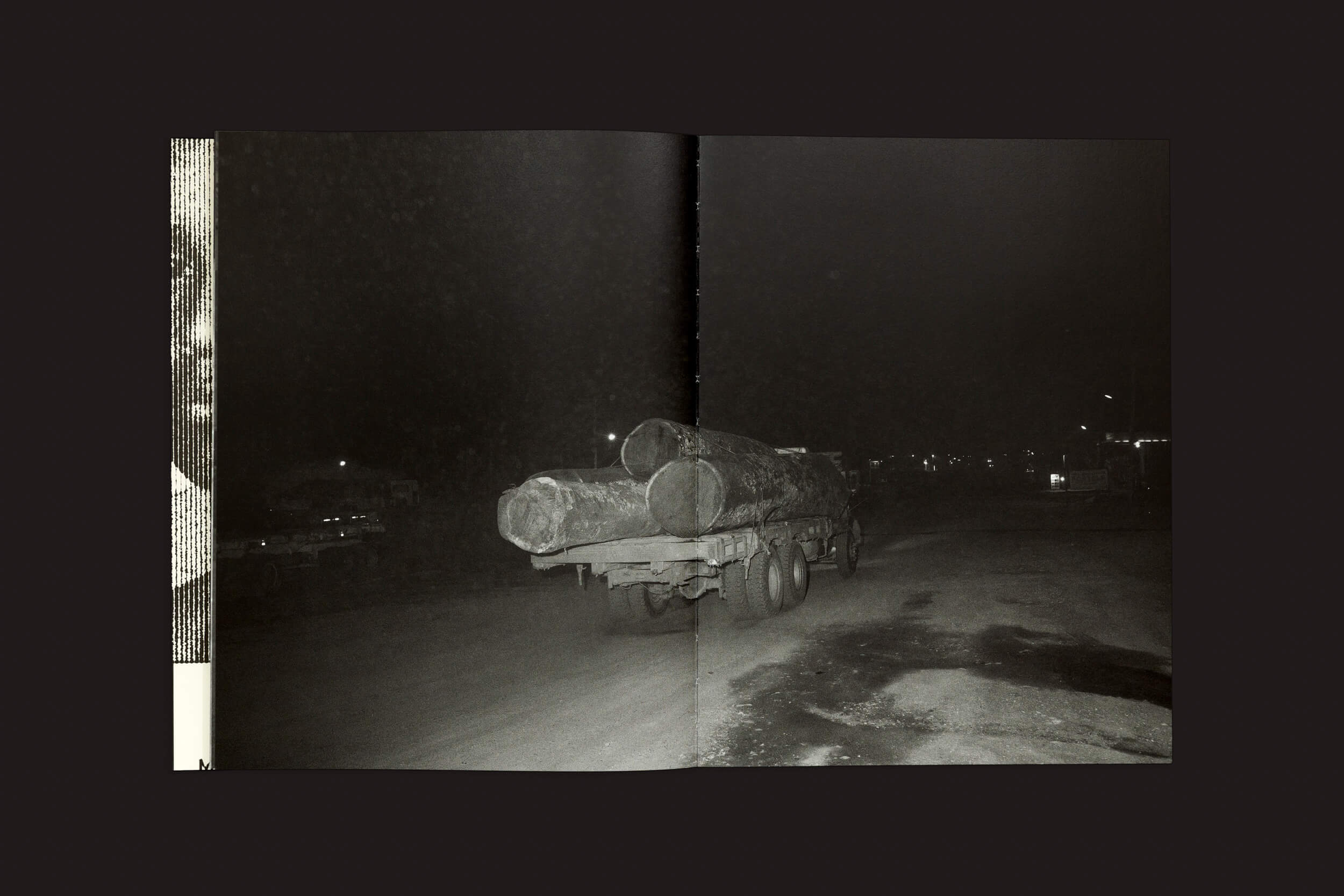A foto noturna de Tommaso Protti: Terra Vermelha de Void mostra um caminhão com grandes toras em uma estrada escura, simbolizando o desmatamento. Central e se afastando, o caminhão é colocado contra luzes fracas à direita, criando uma atmosfera sombria e melancólica.