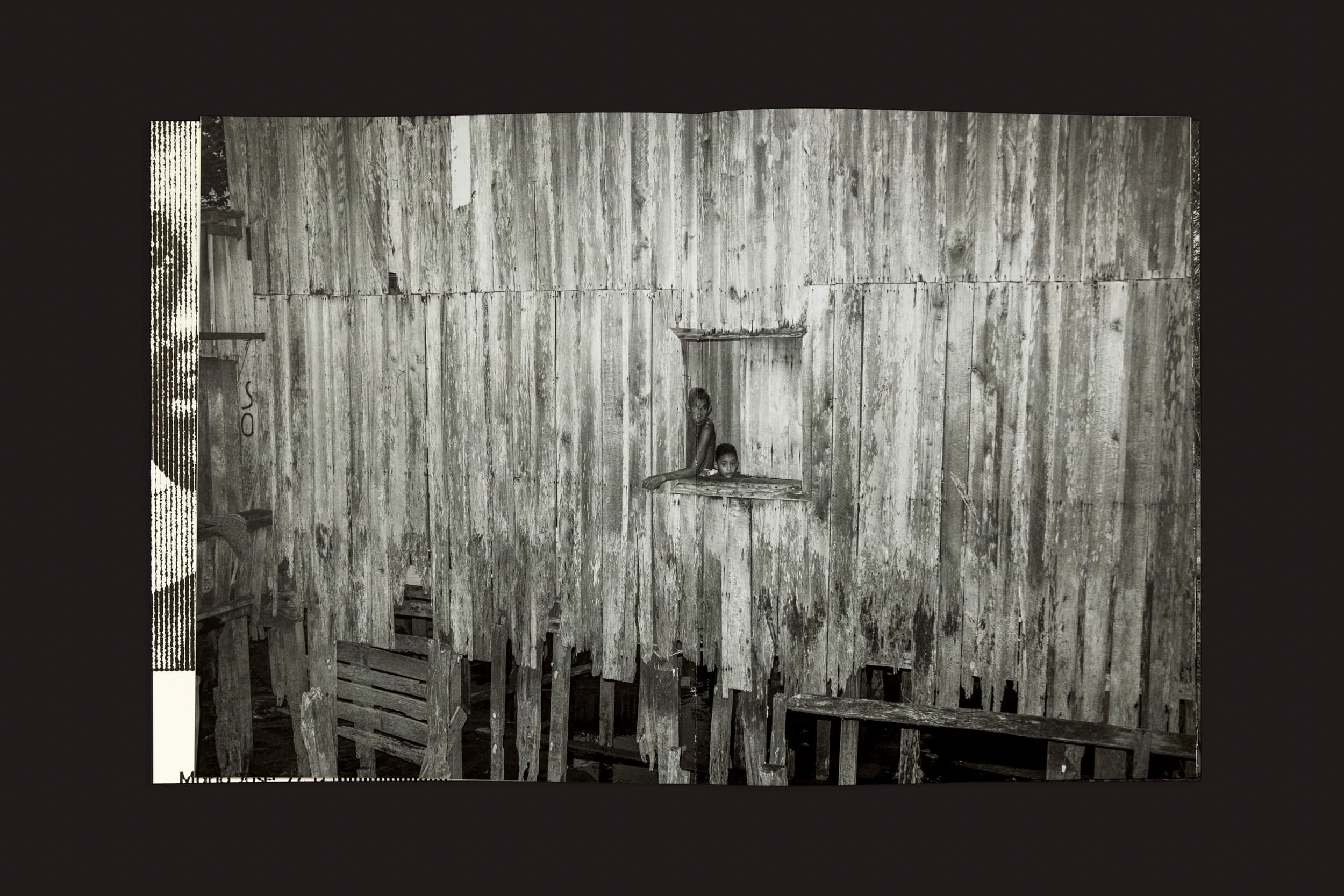 Em Tommaso Protti: Terra Vermelha de Void, uma casa de palafitas de madeira desgastada enfrenta a ameaça do desmatamento. Duas crianças se debruçam em uma janela quadrada, suas expressões curiosas contrastando com a textura envelhecida. O fundo escuro destaca a cena, evocando a urgência pela conservação.