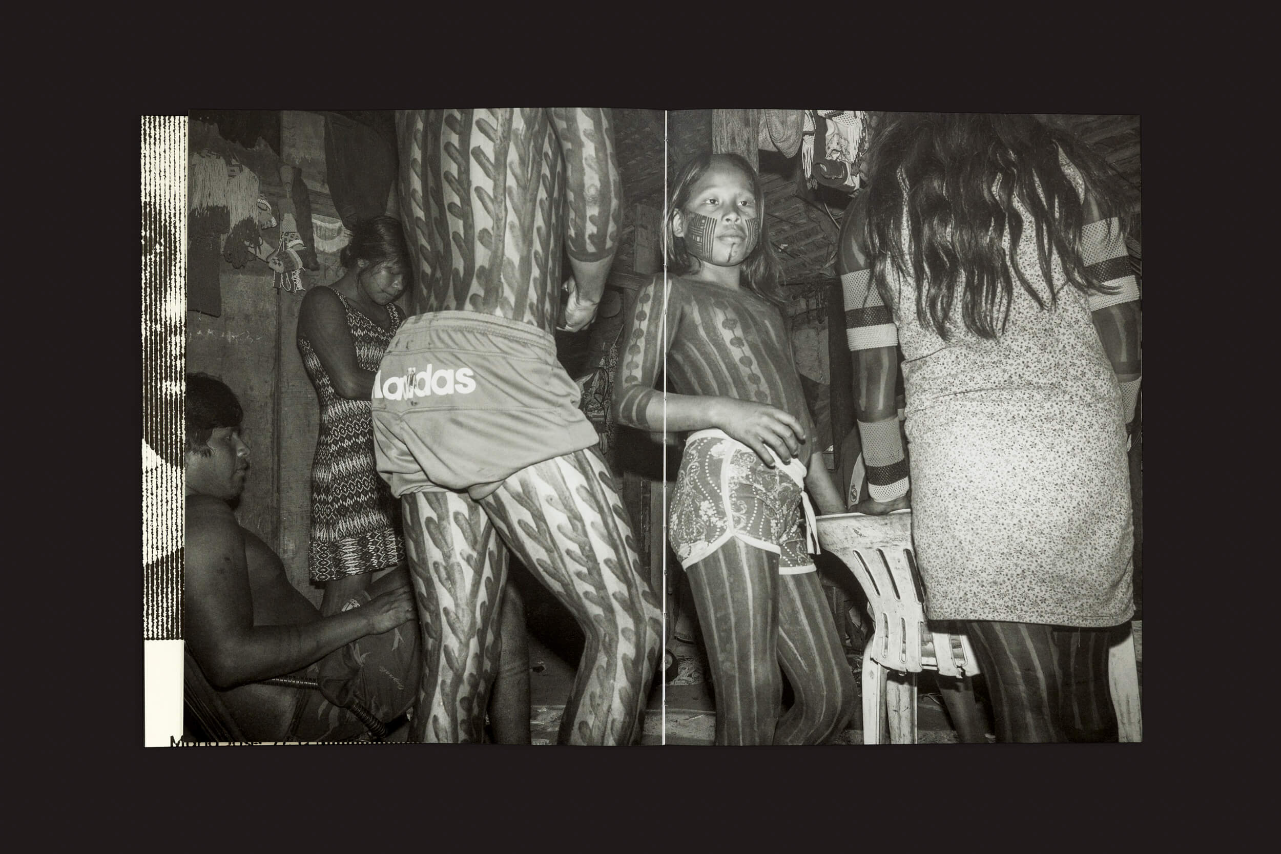 Pessoas com pintura corporal intrincada estão em uma sala, uma usando shorts Adidas e outras em trajes casuais. Sua atmosfera animada e discussões apaixonadas sobre conservação ambiental se misturam perfeitamente com expressões criativas, lembrando temas de Tommaso Protti: Terra Vermelha do Void.