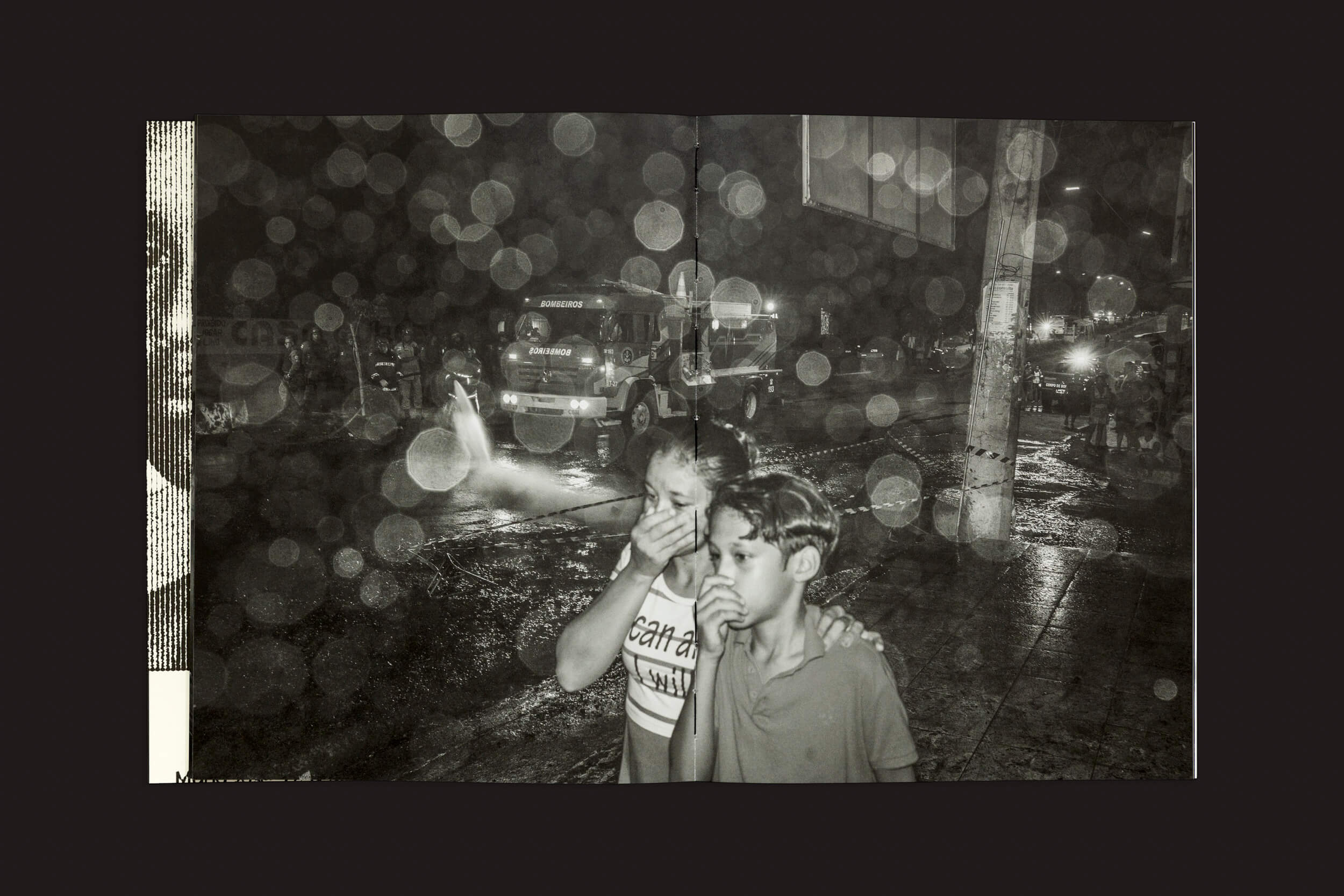 Duas crianças cobrem o nariz em uma rua molhada à noite. Ao fundo, um caminhão de bombeiro e bombeiros jogam água para combater a polícia, com reflexos circulares de luz capturados na imagem intitulada Tommaso Protti: Terra Vermelha por Void.