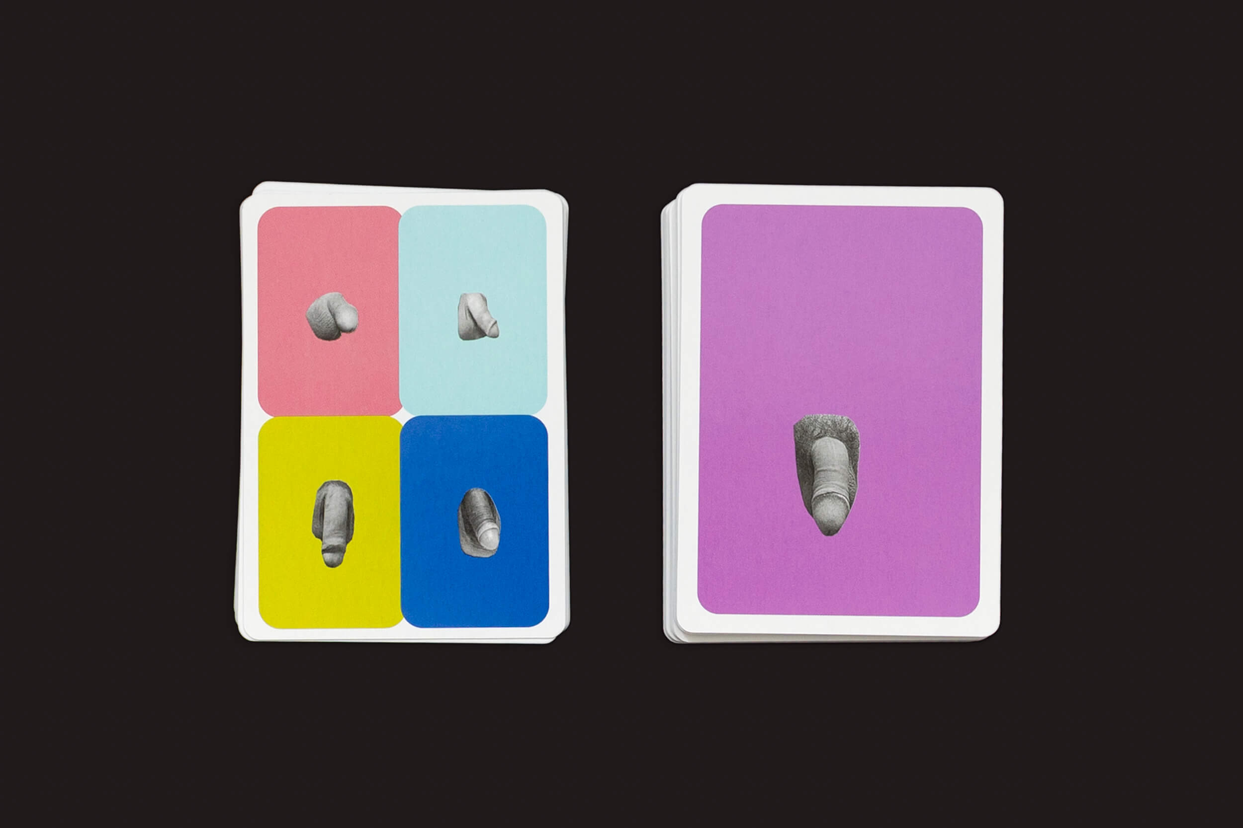 Um baralho de The Lost Greek Penis Game da Fraglich Publishing é mostrado em um fundo preto. Uma carta, separada, tem um fundo roxo com uma imagem de dedo em preto e branco que lembra esculturas coloridas, enquanto o resto exibe seções vibrantes em rosa, ciano, amarelo e azul com motivos de dedos.