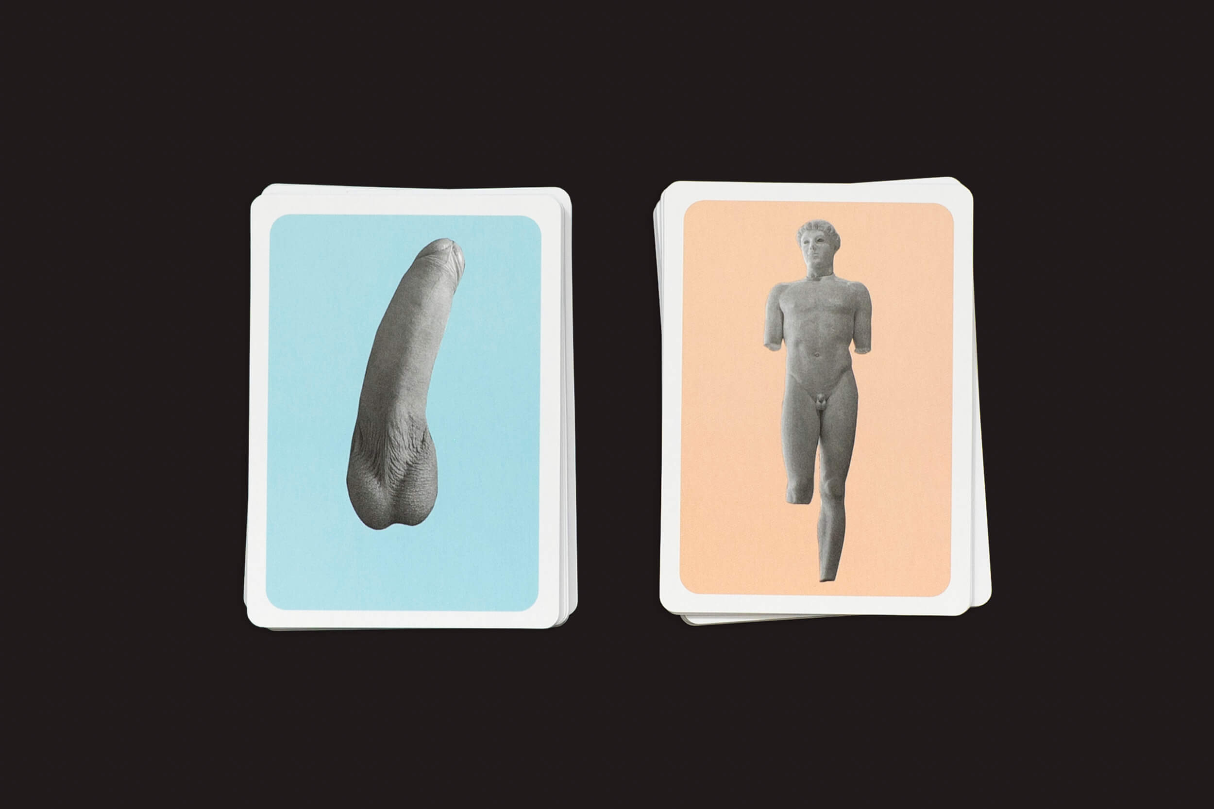 Duas pilhas de cartas contra um fundo escuro; a esquerda mostra um dedo decepado em azul, a direita apresenta uma estátua clássica sem membros em pêssego, reminiscente de esculturas coloridas. De The Lost Greek Penis Game da Fraglich Publishing.