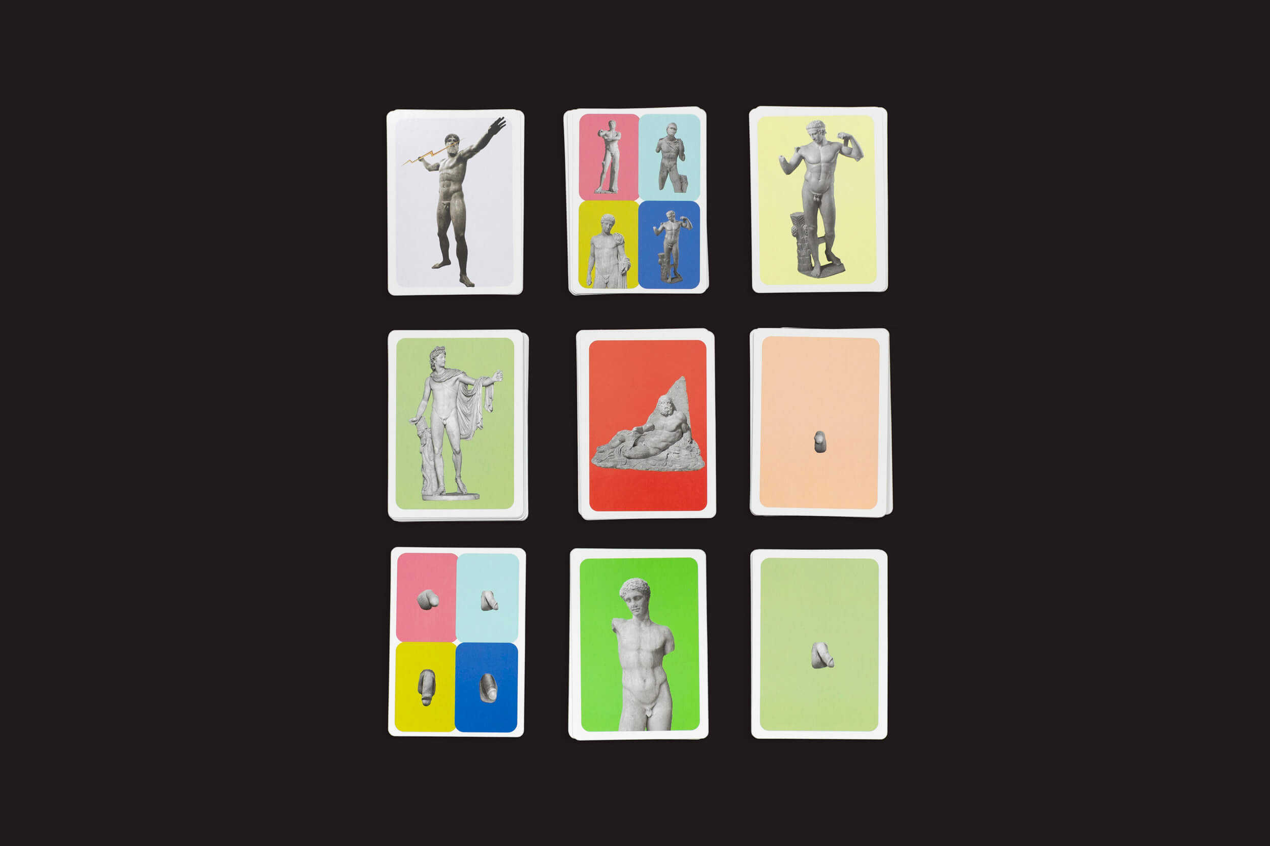 O Jogo do Pênis Grego Perdido da Fraglich Publishing apresenta uma grade de nove cartas em um fundo preto com estátuas e bustos gregos em fundos vibrantes em vermelho, verde e azul, mesclando artisticamente a elegância clássica com contrastes coloridos modernos.