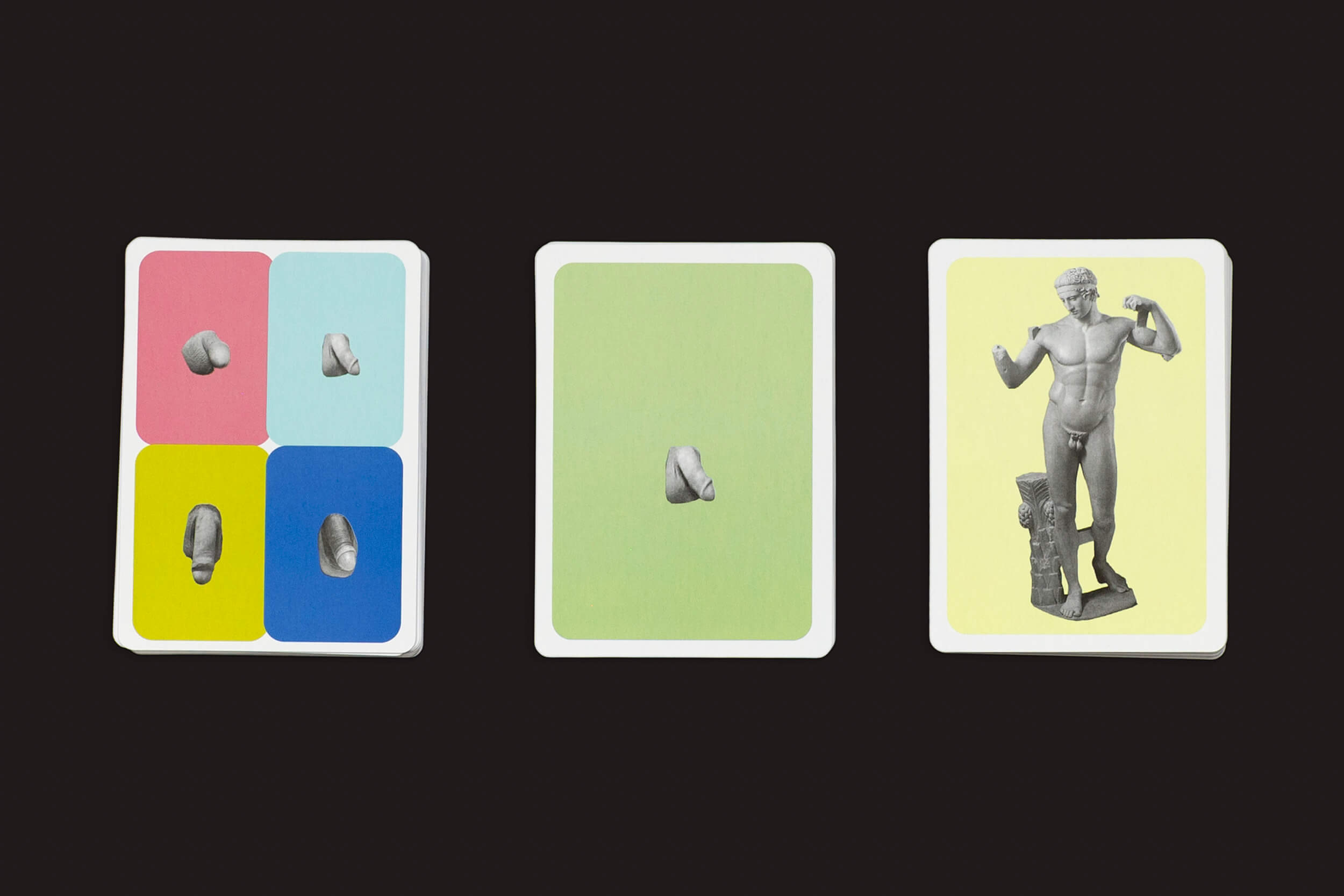 Três cartas de baralho do The Lost Greek Penis Game da Fraglich Publishing aparecem em um fundo preto. A primeira carta lembra esculturas coloridas com quatro seções de diversas partes do corpo, a segunda carta é verde destacando uma única parte do corpo, e a terceira carta apresenta uma estátua grega de uma figura masculina sem um membro.