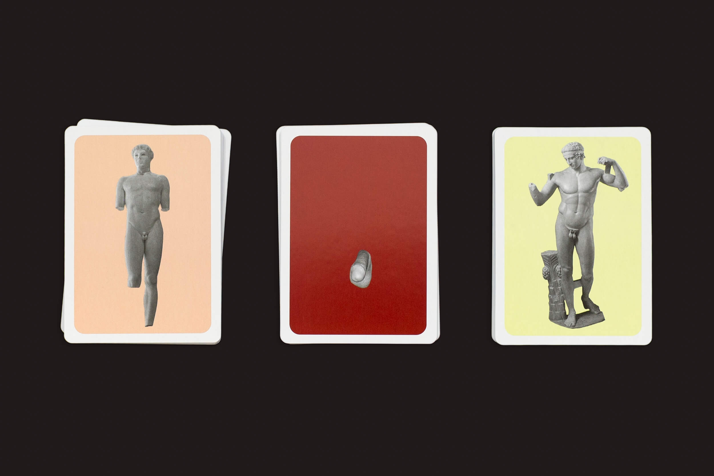 Três cartas de The Lost Greek Penis Game da Fraglich Publishing estão sobre um fundo escuro: a carta da esquerda mostra uma estátua em pé, a do meio revela um fragmento de pedra e a da direita retrata uma estátua flexionada ao lado de um pedestal. Fundos evocativos acentuam o tema das estátuas gregas.
