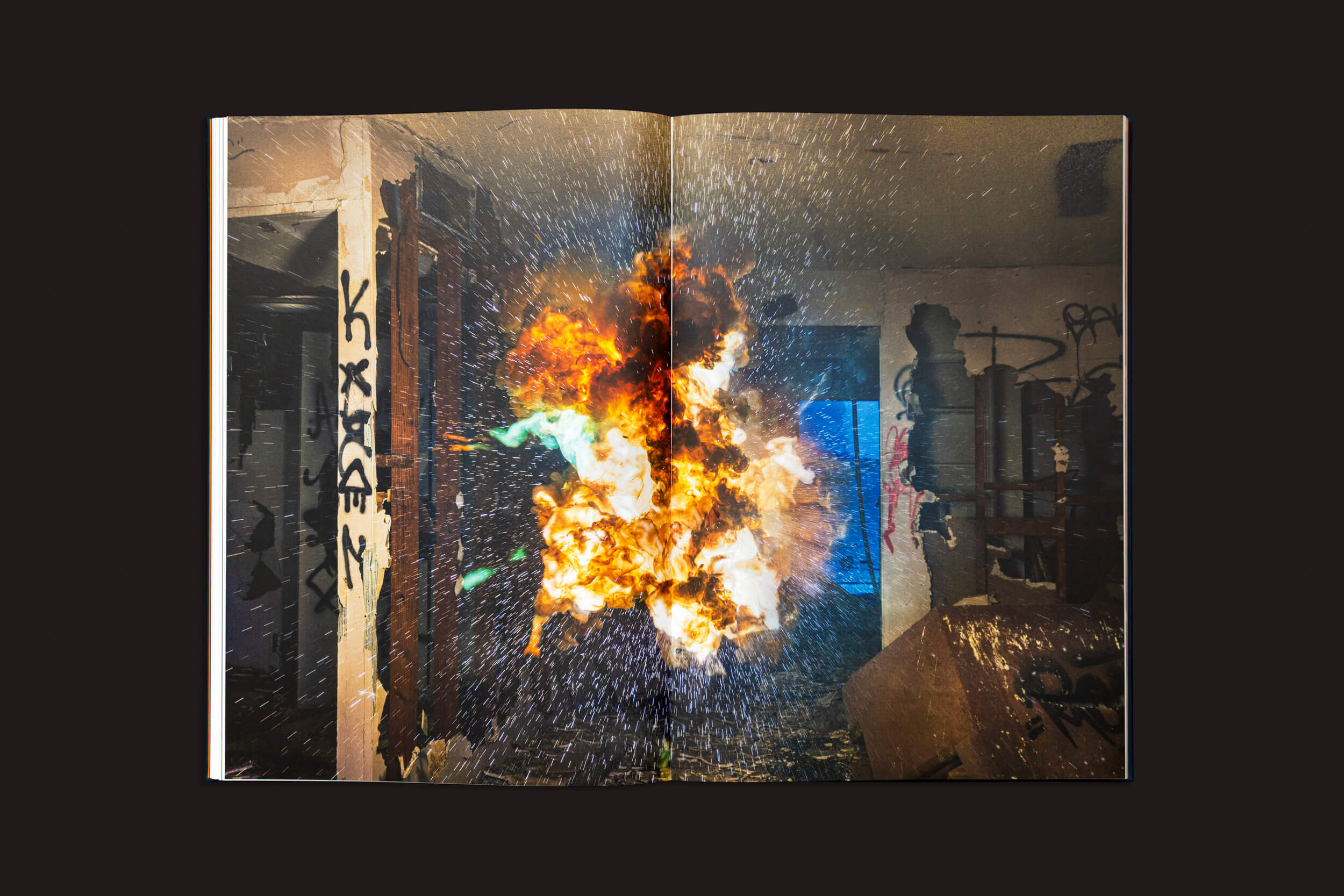 Em "Kevin Cooley: The Wizard of Awe", de The Eriskay Connection, um livro aberto mostra uma explosão vívida com chamas e destroços em uma sala escura, contraposta a paredes cobertas de Pirotécnico e fragmentos espalhados, realçando o caos dramático contra o fundo preto.