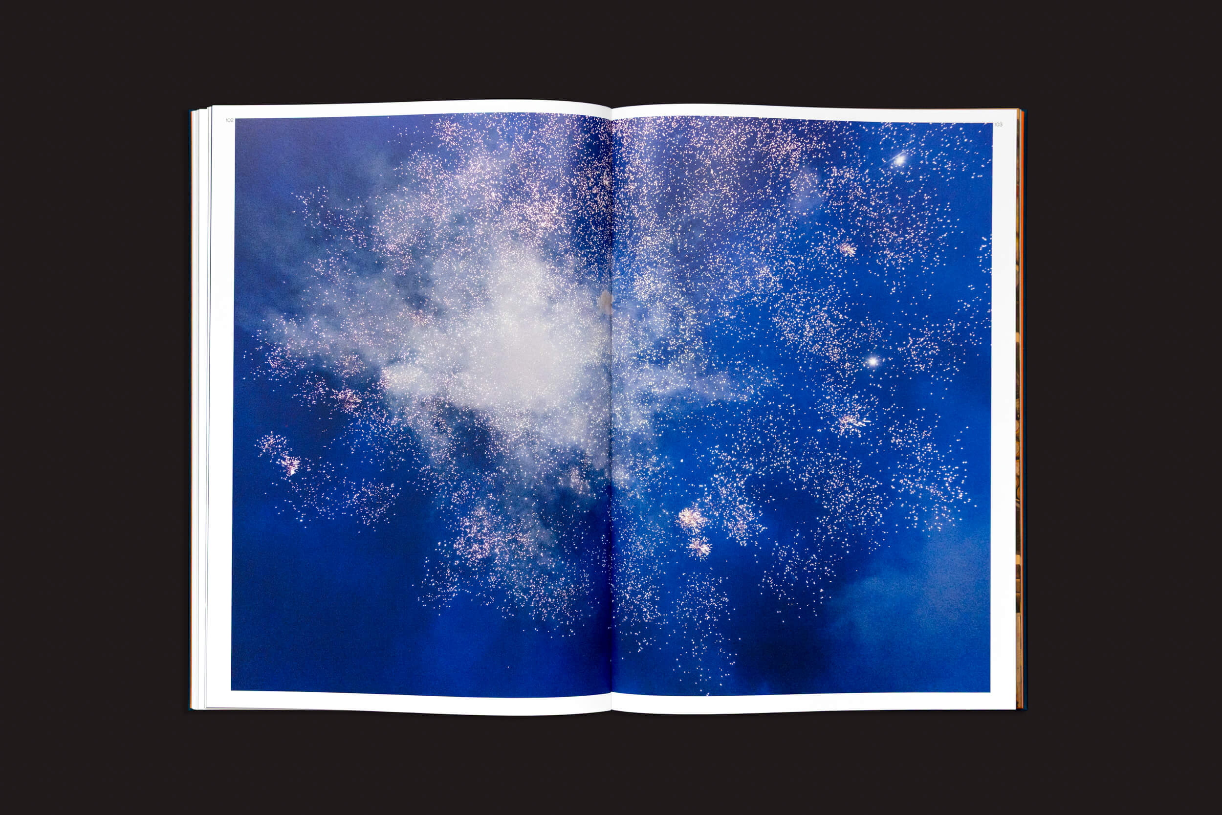 Em "Kevin Cooley: The Wizard of Awe", da The Eriskay Connection, uma fotografia apresenta brilhos brilhantes, semelhantes a estrelas, contra um fundo azul-escuro, que lembra uma galáxia ou fogos de artifício. A imagem ocupa ambas as páginas, criando uma cena celestial coesa e vibrante, com um encanto quase pirotécnico.