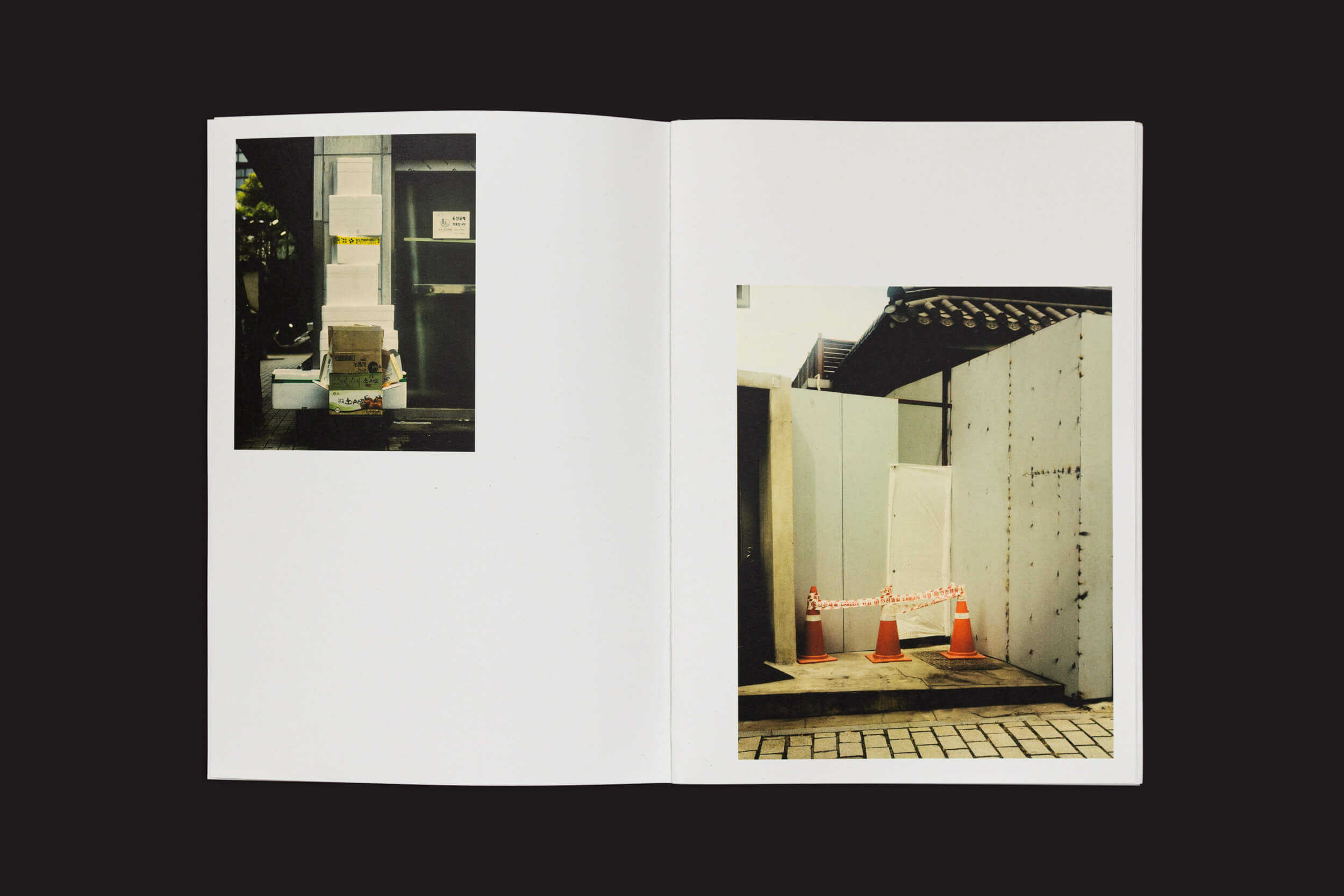 As páginas abertas de Eduard Sánchez Ribot: Tierra, Camino, Fruto, da Handshake, revelam uma pilha de caixas perto de uma porta à esquerda e uma zona de construção com cones e barreiras de fita à direita, tudo colocado de forma pungente contra um fundo preto.