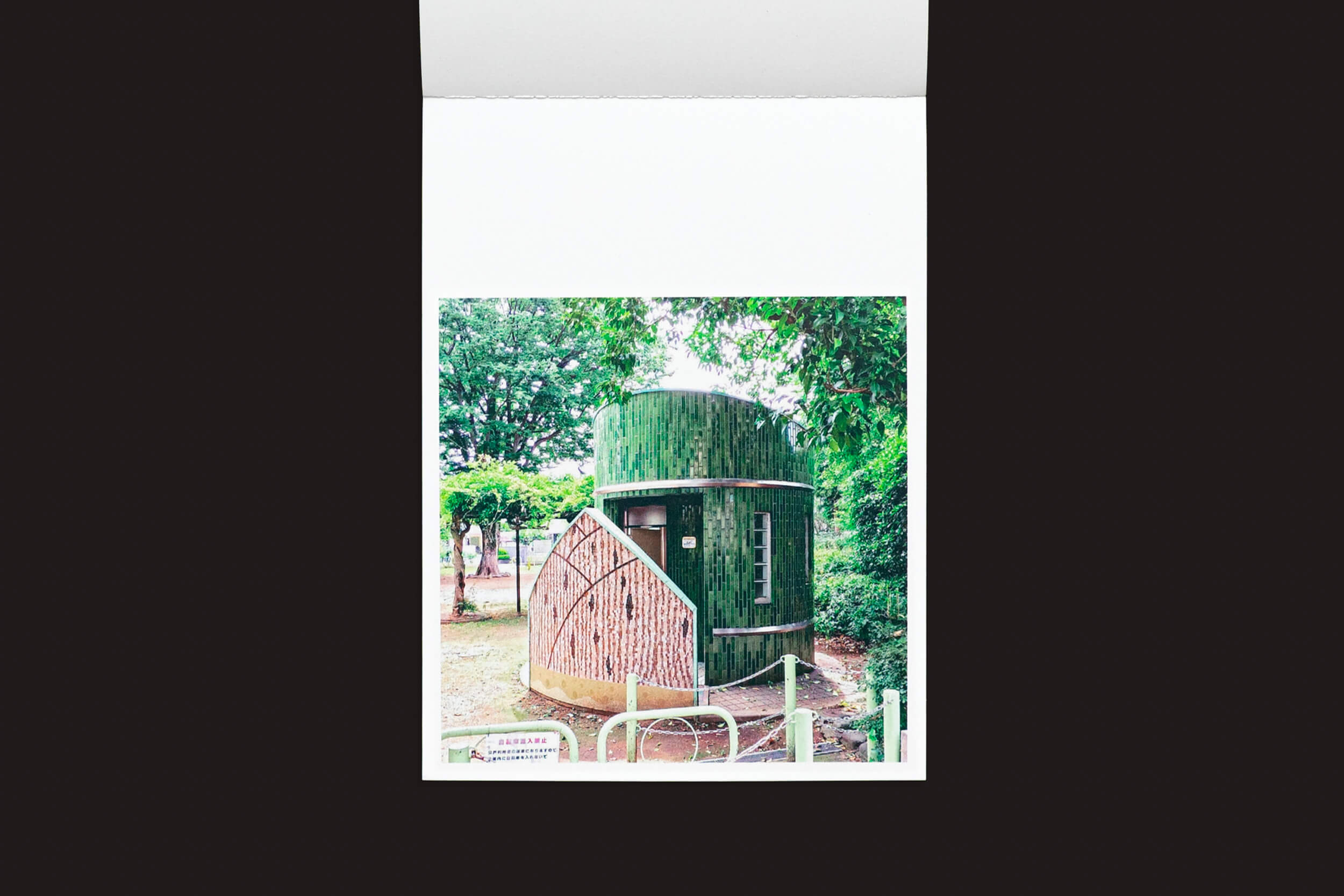 Em um parque, um pequeno edifício cilíndrico verde, inspirado na arquitetura japonesa e apresentando um painel com padrão de madeira e telhado verde, é capturado em Hidefumi Nakamura: Toilets a Go Go! por Handshake. Um caminho gracioso leva à sua entrada em meio às árvores ao redor.