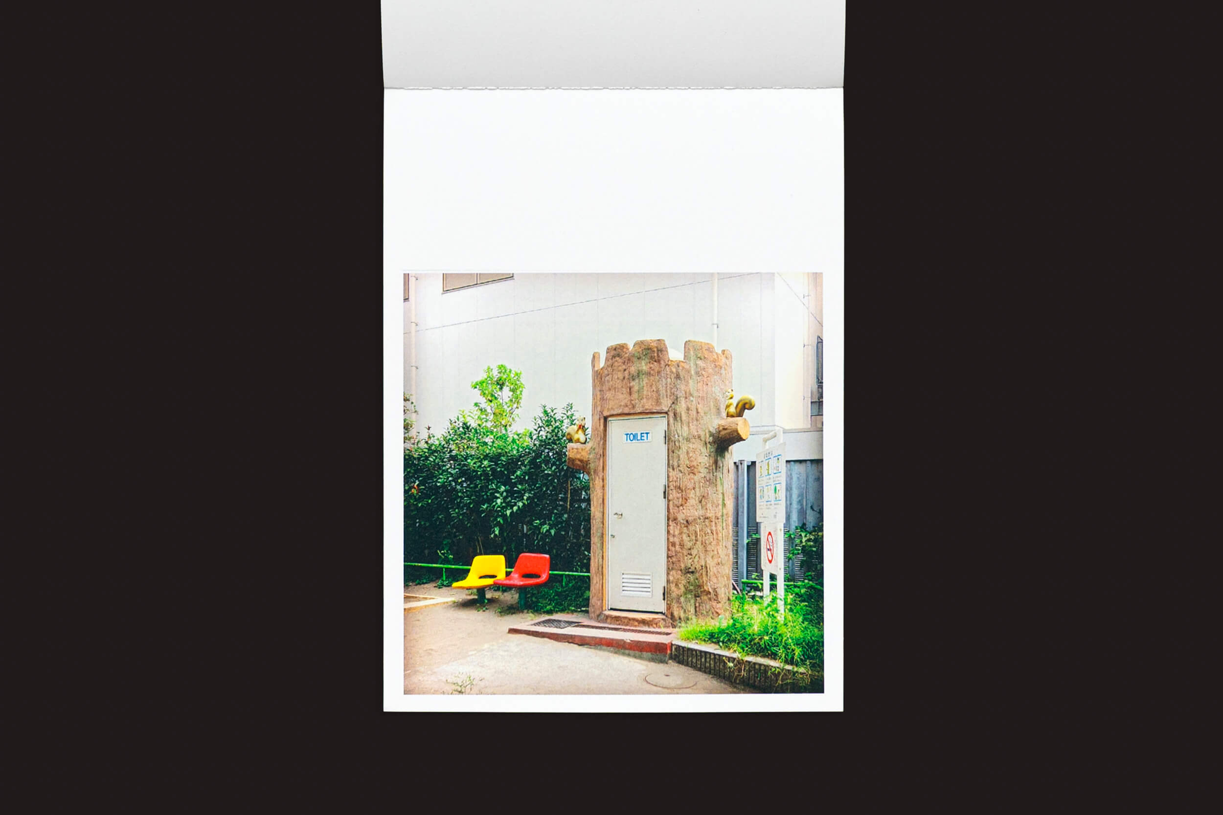 Uma foto captura um banheiro público em formato de castelo com a porta central marcada como TOILET, cercado por cadeiras de plástico vermelhas e amarelas vibrantes. Aninhado em vegetação, ele combina design caprichoso com praticidade, ecoando o estilo inovador apresentado em Hidefumi Nakamura: Toilets a Go Go! da Handshake.