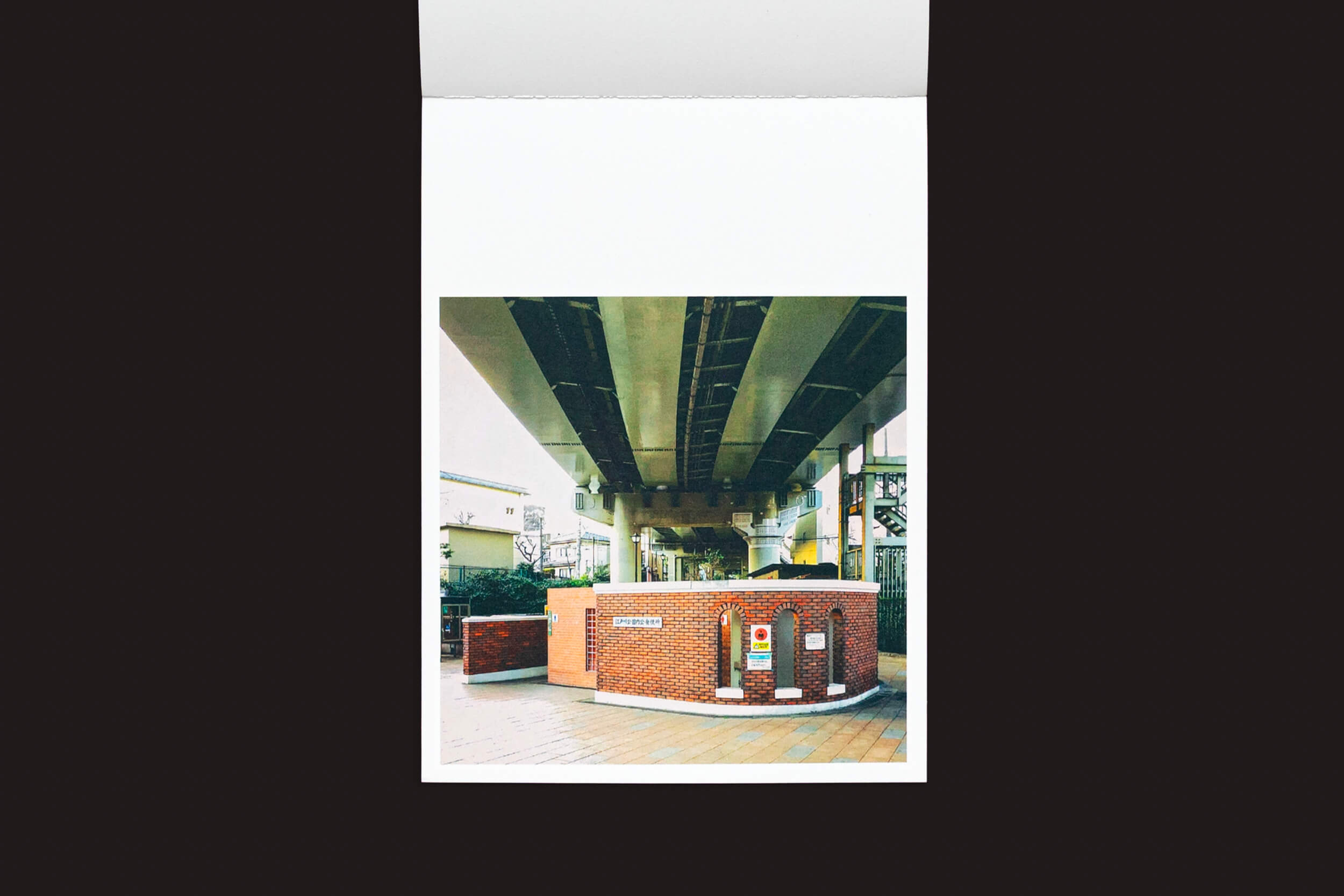 Foto de Hidefumi Nakamura: Toilets a Go Go! contra um fundo escuro, mostrando uma estrutura de tijolos vermelhos com uma máquina de venda automática abaixo de uma estrada elevada. A cena, com elementos de arquitetura japonesa de Handshake, inclui uma passarela e prédios próximos.