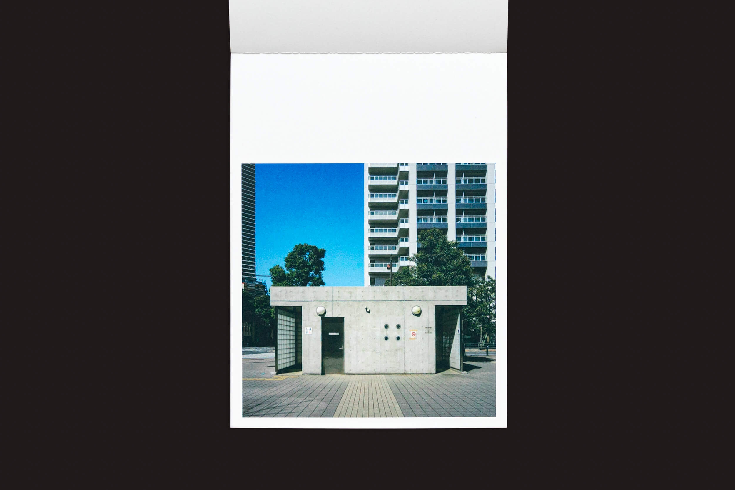 Um instantâneo de Hidefumi Nakamura: Toilets a Go Go! da Handshake mostra um banheiro minimalista com uma fachada de concreto, ecoando a arquitetura japonesa em um ambiente urbano. Prédios altos e árvores se destacam contra um fundo de céu azul claro.