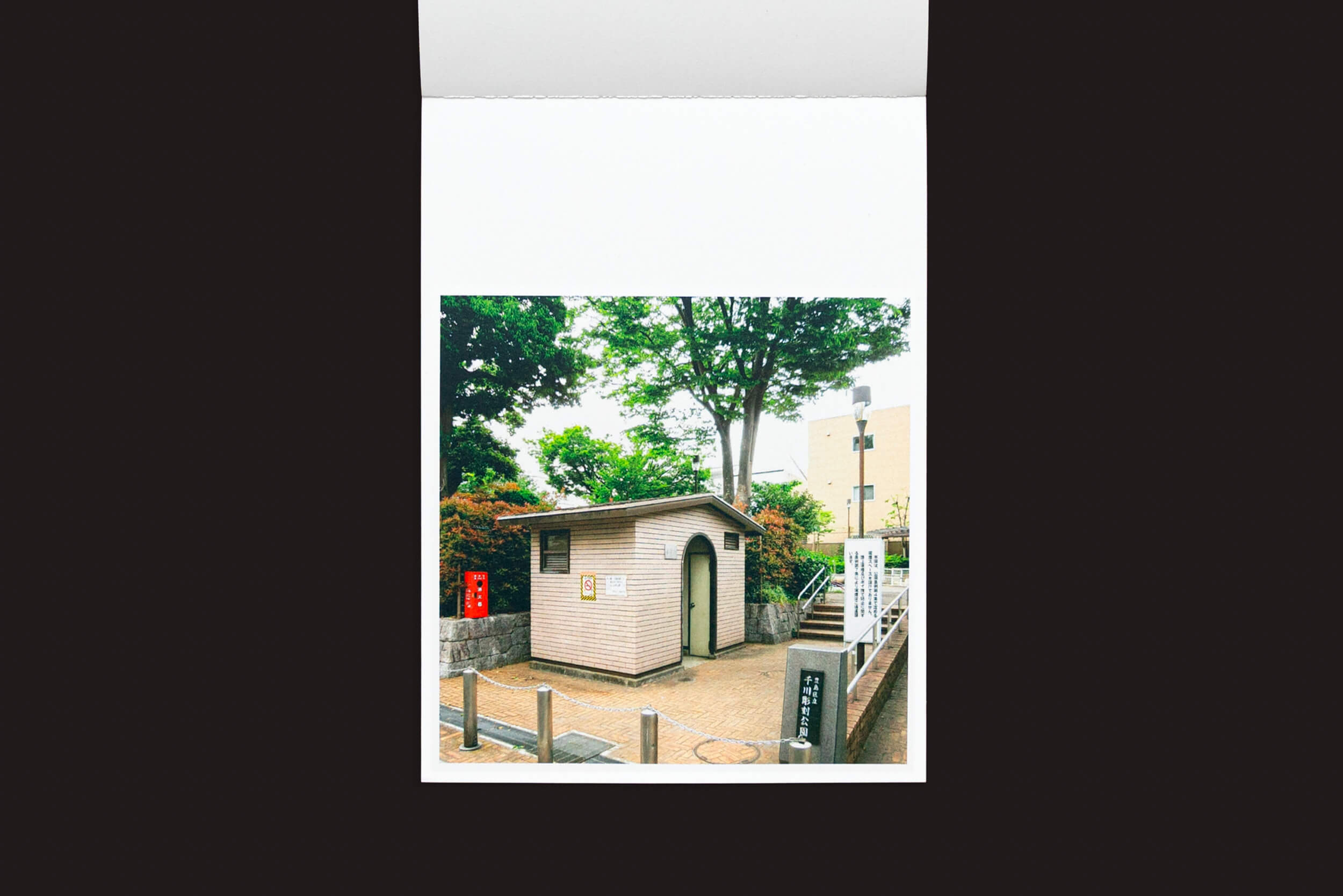 Aninhado em meio à vegetação, um edifício de um andar com telhado de telhas e porta em arco reflete a arquitetura japonesa tradicional. Cercado por árvores, arbustos, uma grade branca e postes sob um céu brilhante, é ideal para capturar com Hidefumi Nakamura: Toilets a Go Go! por Handshake.