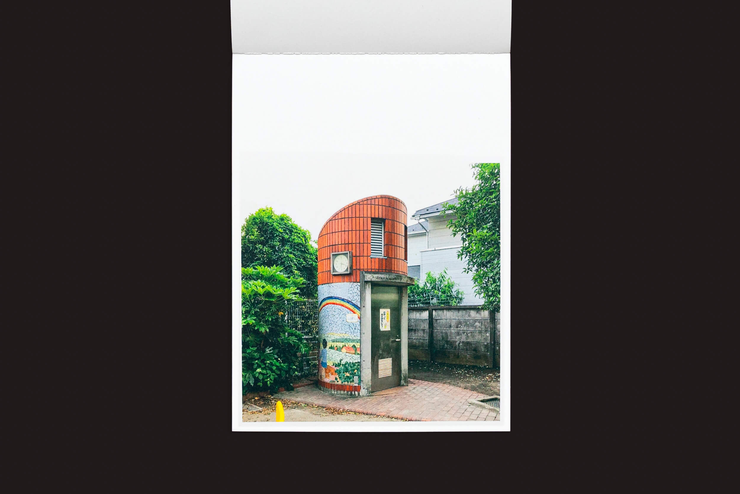 Um pequeno estande de Hidefumi Nakamura: Toilets a Go Go! da Handshake apresenta um telhado de telhas e um mural colorido inspirado na arquitetura japonesa em um caminho de tijolos, cercado por vegetação, adicionando um toque artístico, com um edifício visível ao fundo.