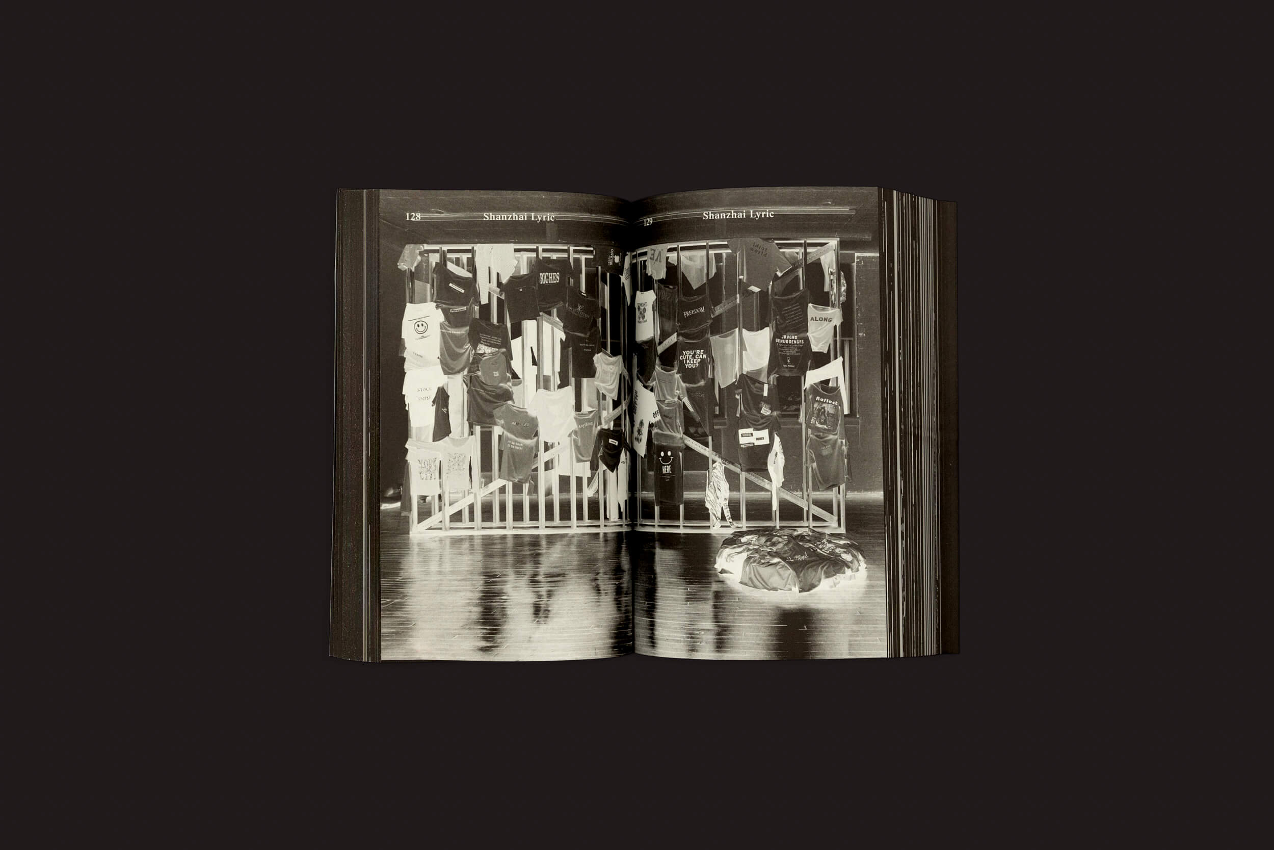 O livro aberto monocromático "UNLICENSED – Bootlegging As a Creative Practice: Ben Schwartz" de Valiz mostra uma foto memorial com camisas e bandeiras em uma cerca, objetos no chão e páginas escuras contrastando com o fundo preto.