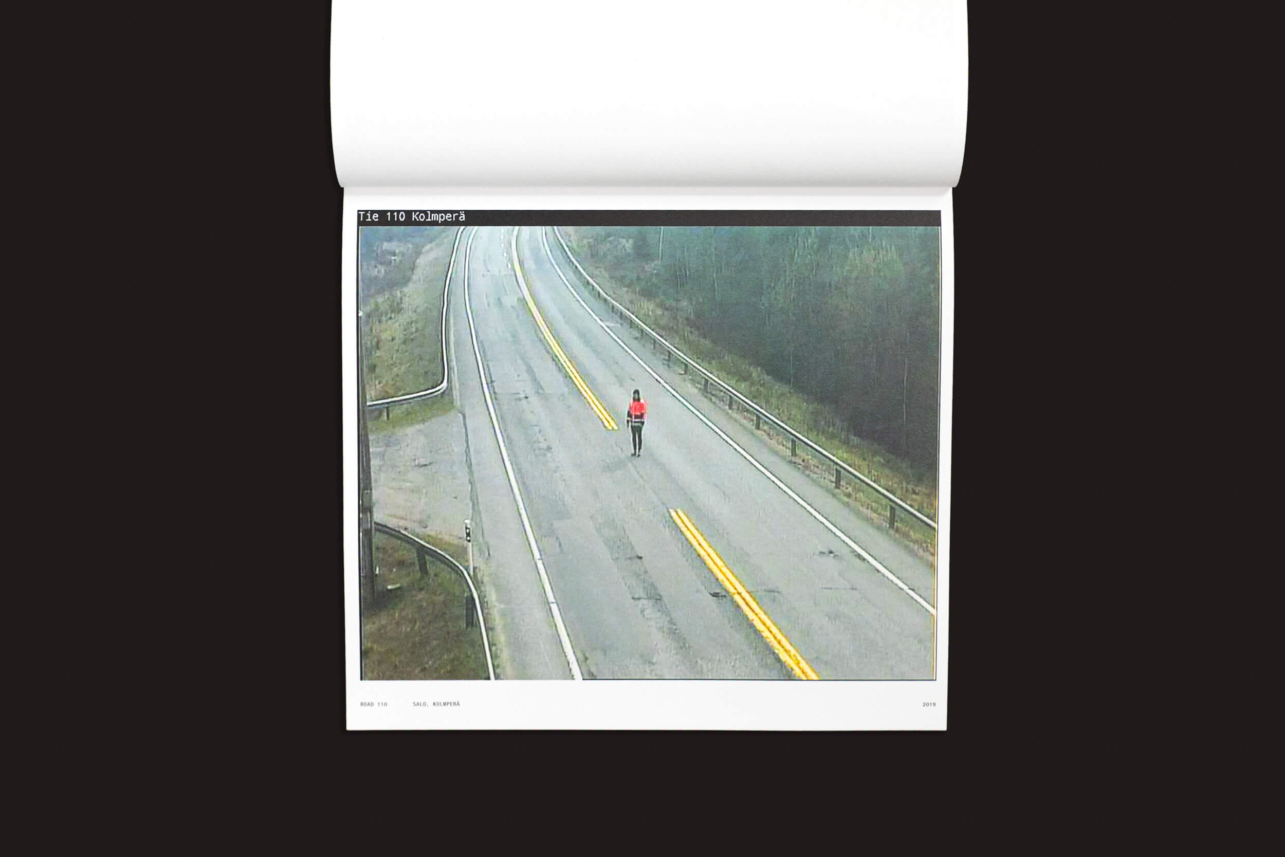 Uma vista aérea mostra uma pessoa com uma jaqueta vermelha, semelhante ao estilo de Tatu Gustafsson em "I on the Road / Weather Camera Self-Portraits, 2012-2021", da Fw:Books, caminhando por uma estrada sinuosa e vazia com linhas amarelas duplas, cercada por grama e árvores sob um céu nublado.